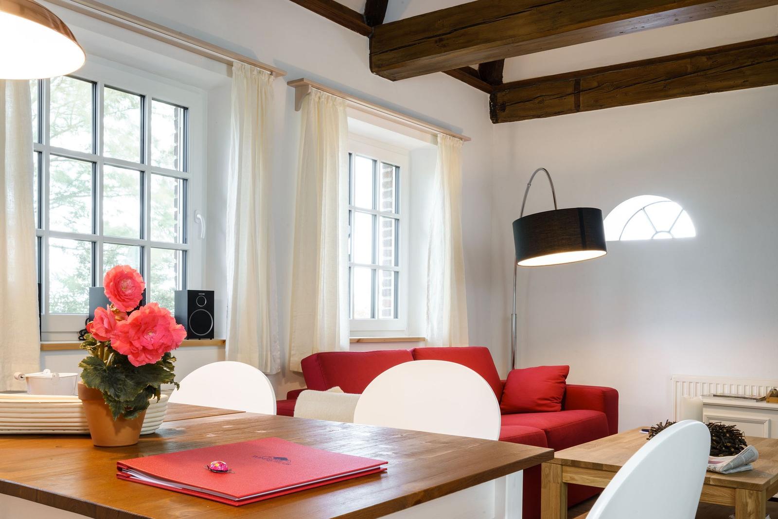 Ein Wohnzimmer mit rotem Sofa, Holztisch und Fenstern mit weißen Vorhängen.