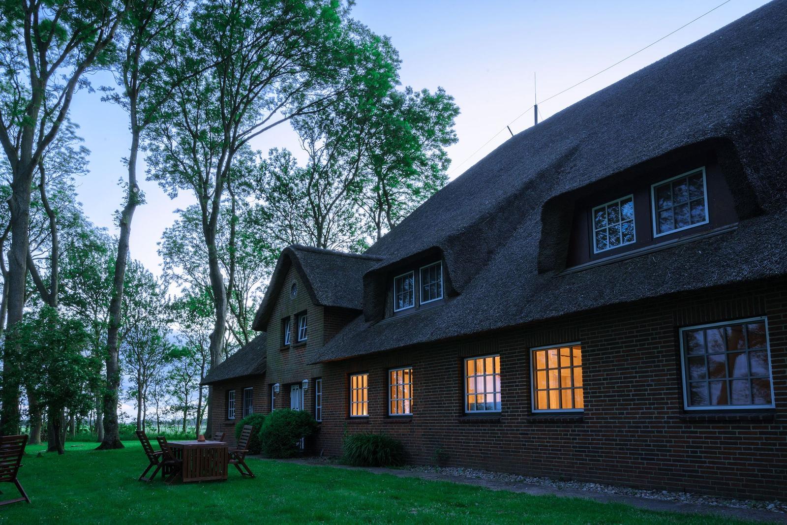 Haus mit Strohdach und erleuchteten Fenstern, umgeben von Bäumen und Gras.