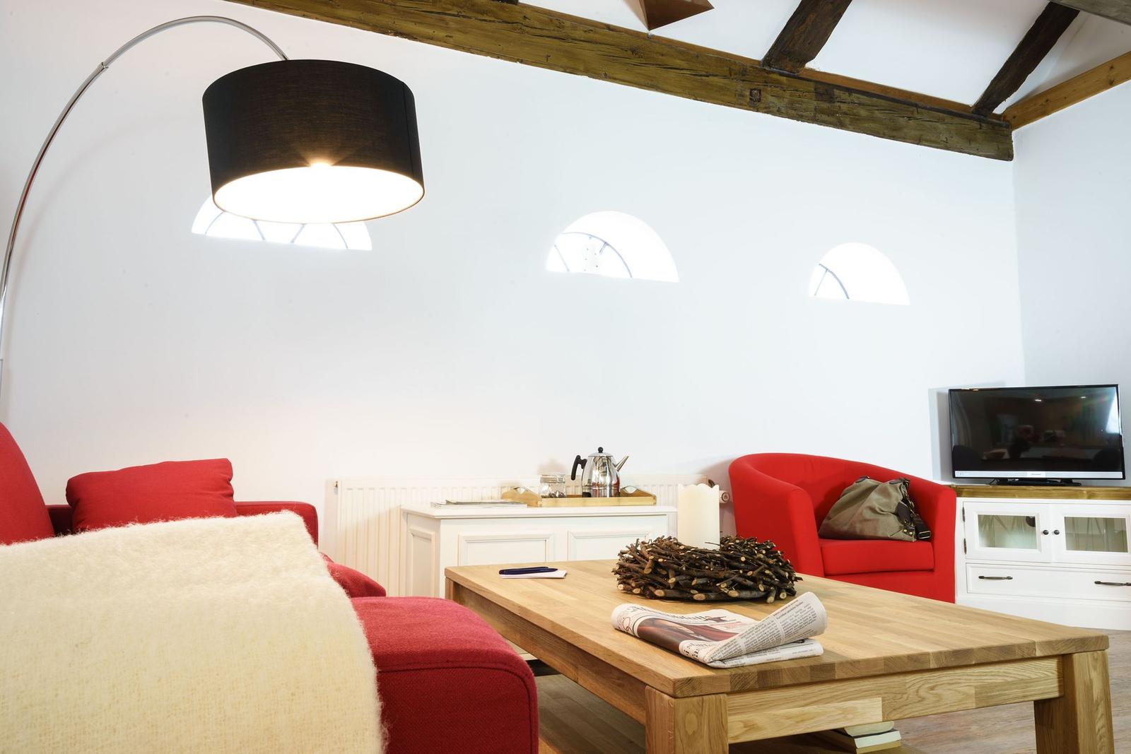 Wohnzimmer mit roten Sesseln, Holztisch und Balkendecke. Drei Bogenfenster und Stehlampe.