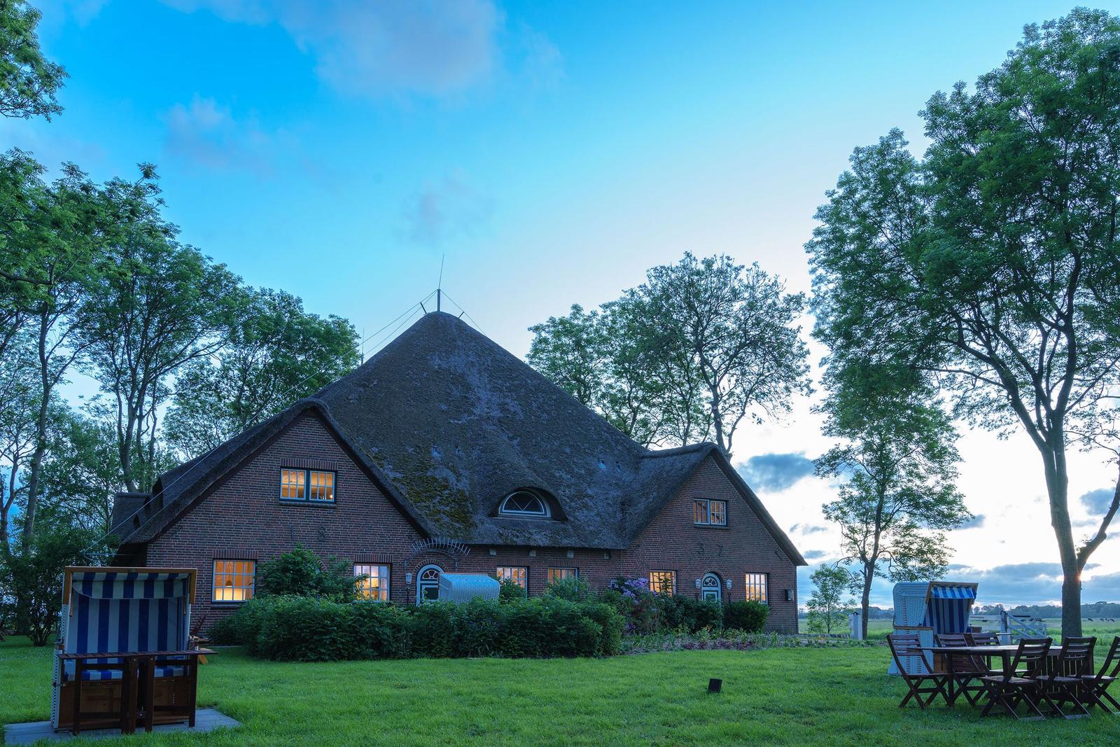 Haus mit strohgedecktem Dach, umgeben von Bäumen und Garten mit Liegestühlen.