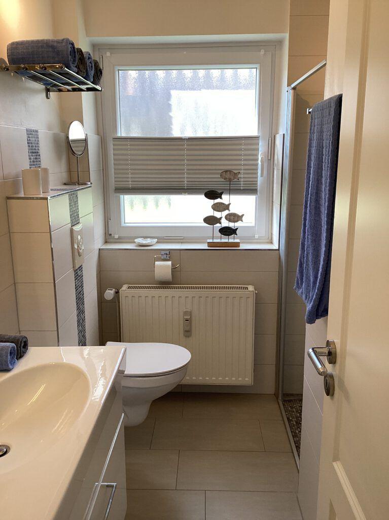 Badezimmer mit Toilette, Waschbecken und Dusche. Fenster mit Jalousie und Badewannenrand.