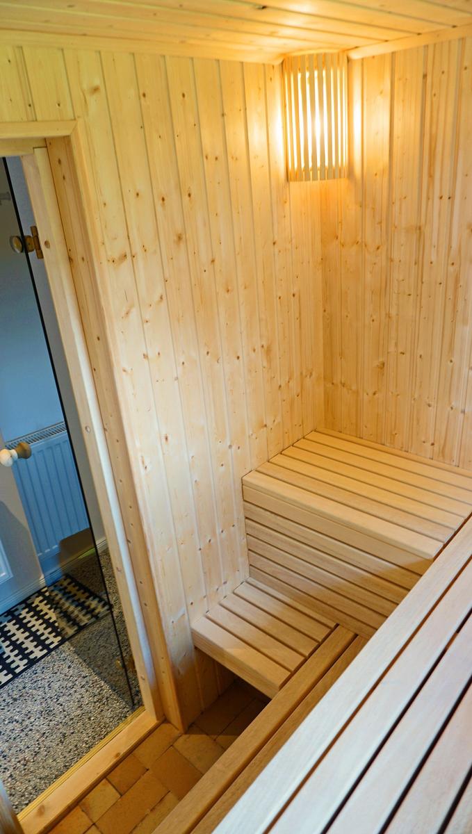 Sauna mit Holzwand und Sitzbänken, beleuchtet durch Wandlampe, Tür zum angrenzenden Raum.