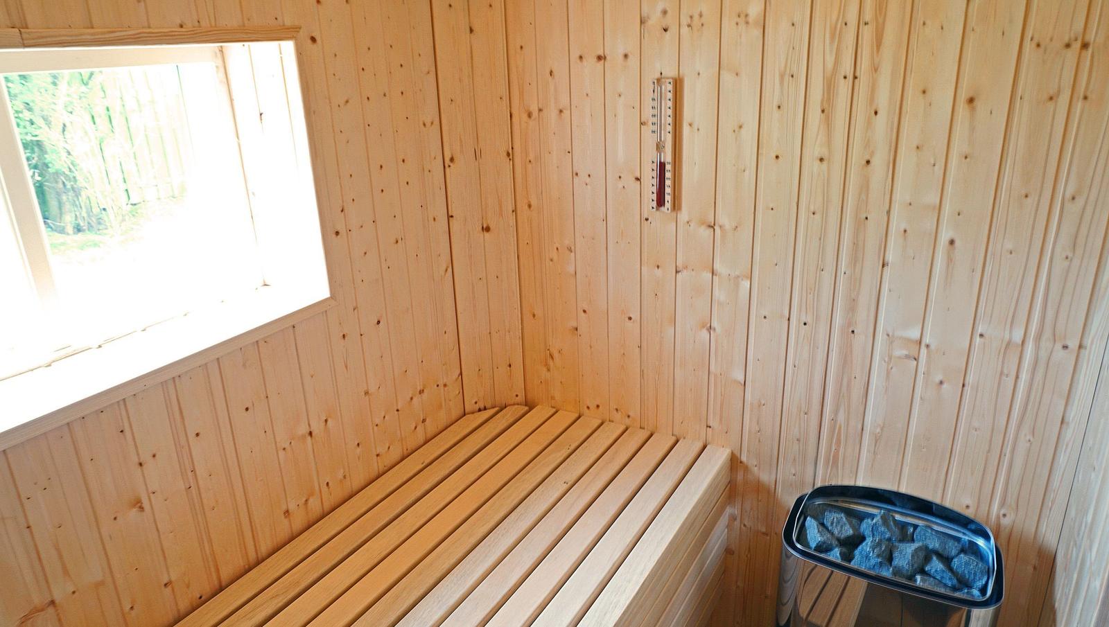Sauna mit Holzwand, Sitzbank und Steinbehälter. Fenster zeigt grüne Landschaft.