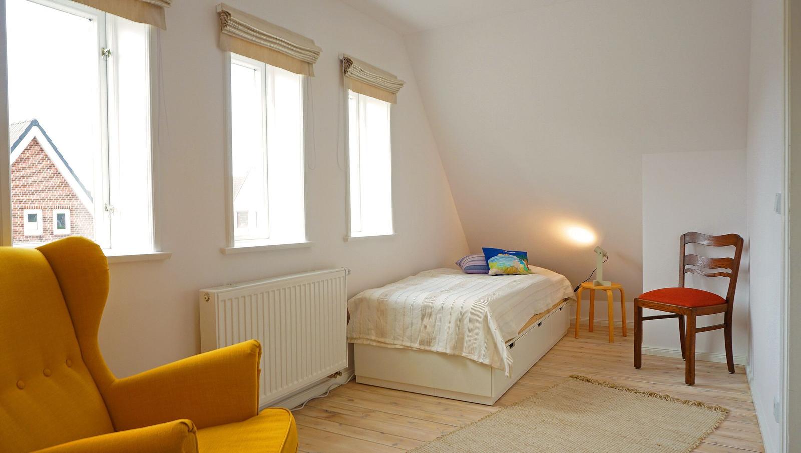 Schlafzimmer mit Bett, Stuhl, Sessel und drei Fenstern. Holzboden und Heizkörper sichtbar.
