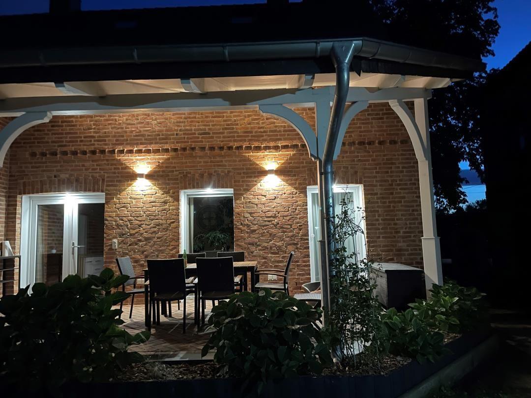 Terrasse mit Tisch und Stühlen unter einem Dach, beleuchtet, vor einer Backsteinwand.