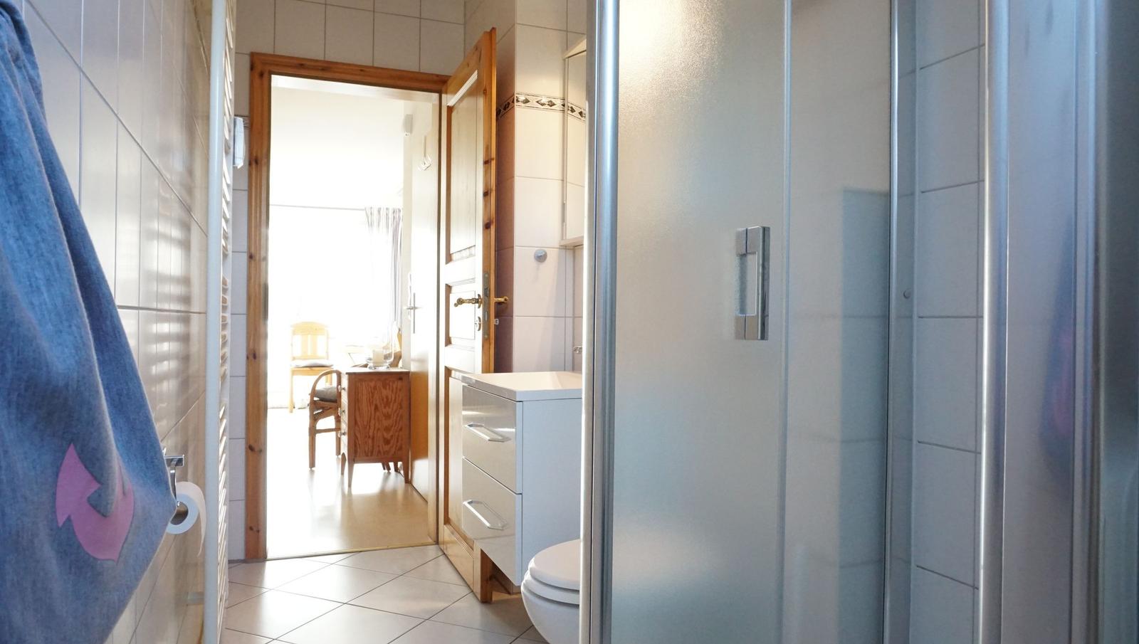 Badezimmer mit Dusche, Toilette und Waschbecken. Durchgang zu einem Raum mit Holztür.