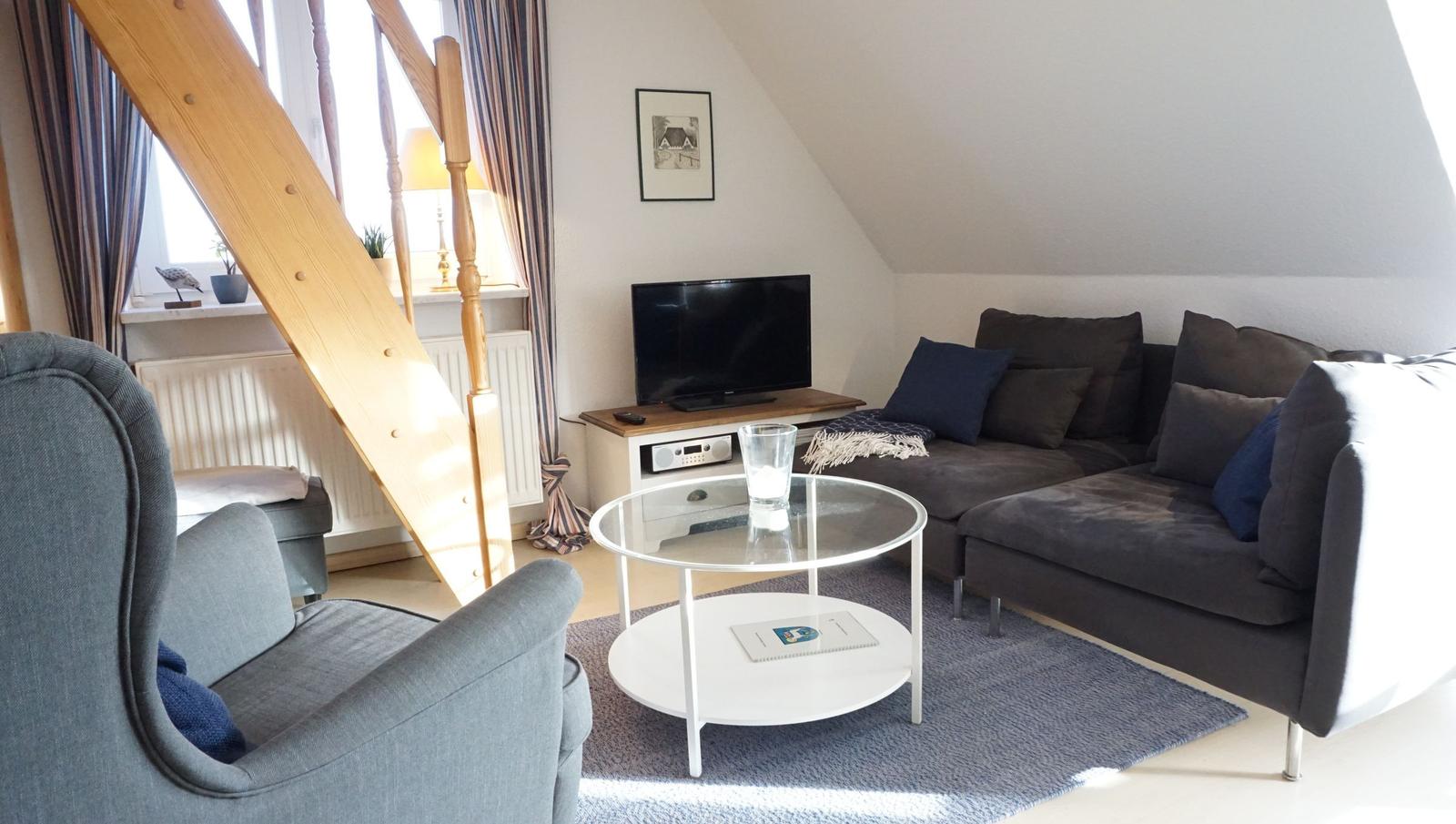 Wohnzimmer mit Sofa, Sessel, Glas-Tisch und Fernseher unter schrägem Dach