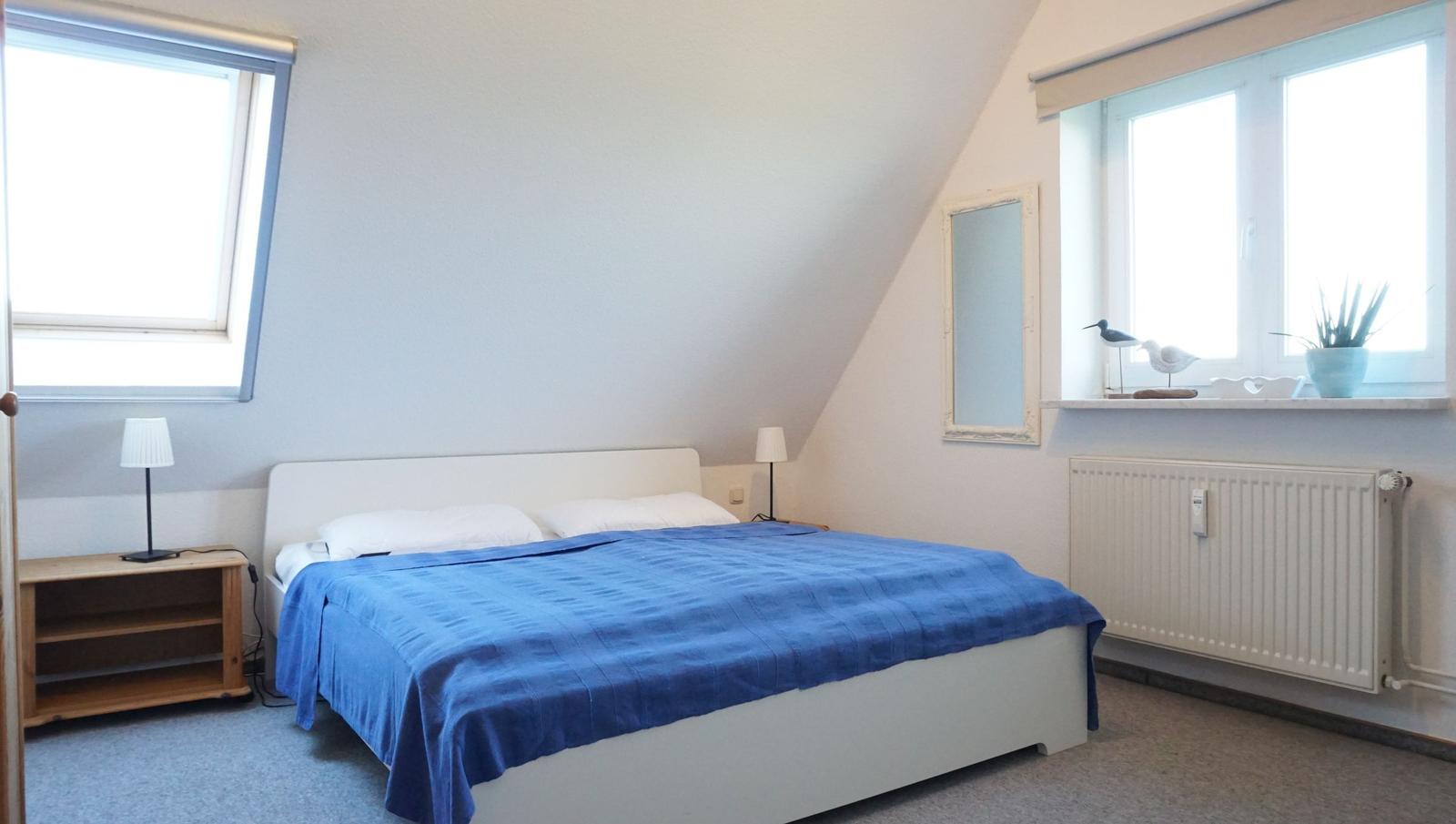 Schlafzimmer mit Dachschrägen, Bett mit blauer Bettwäsche, zwei Fenster und Spiegel an der Wand.