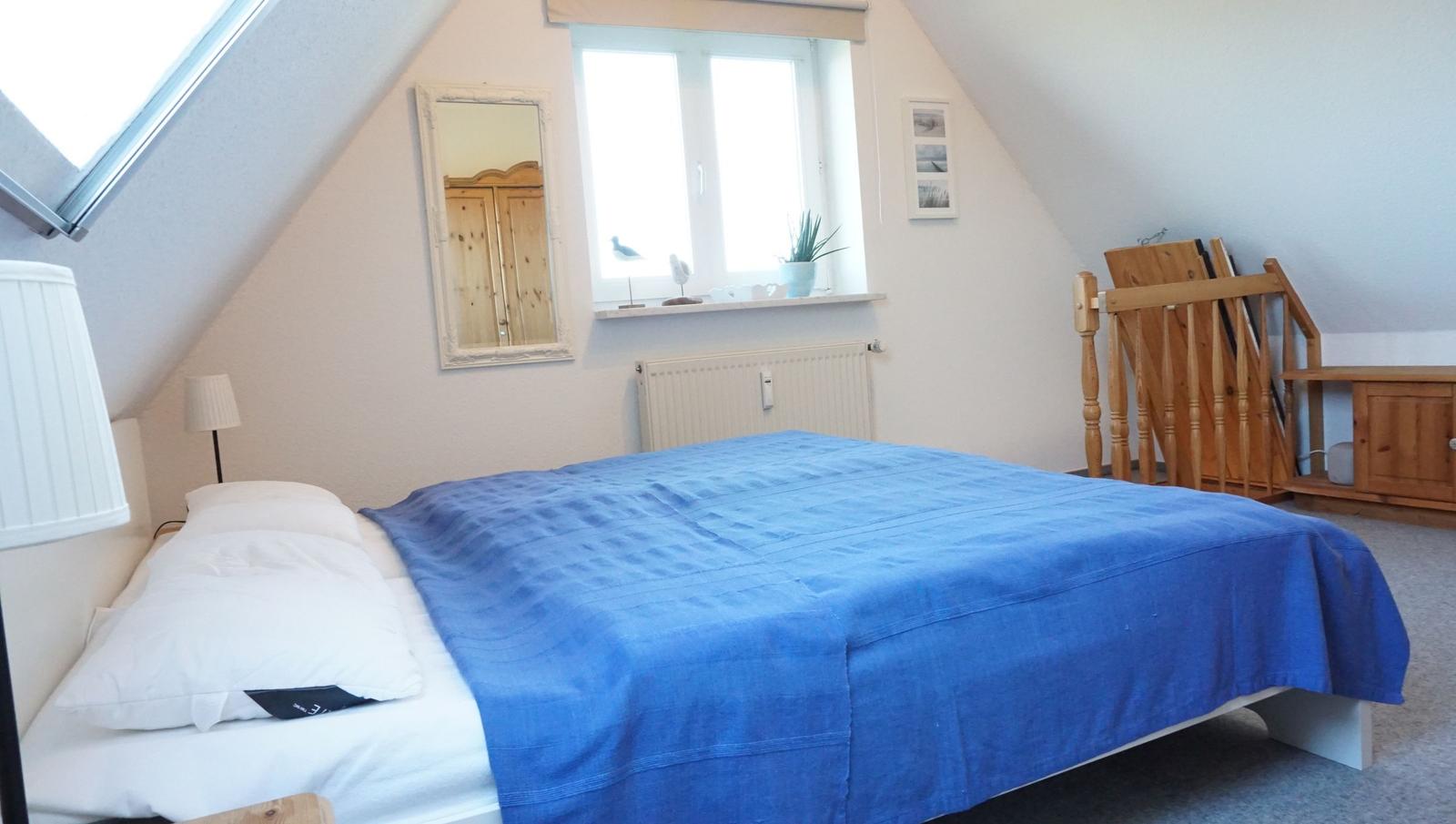Schlafzimmer mit Dachschrägen, Bett mit blauer Bettdecke, Fenster und Spiegel an der Wand.