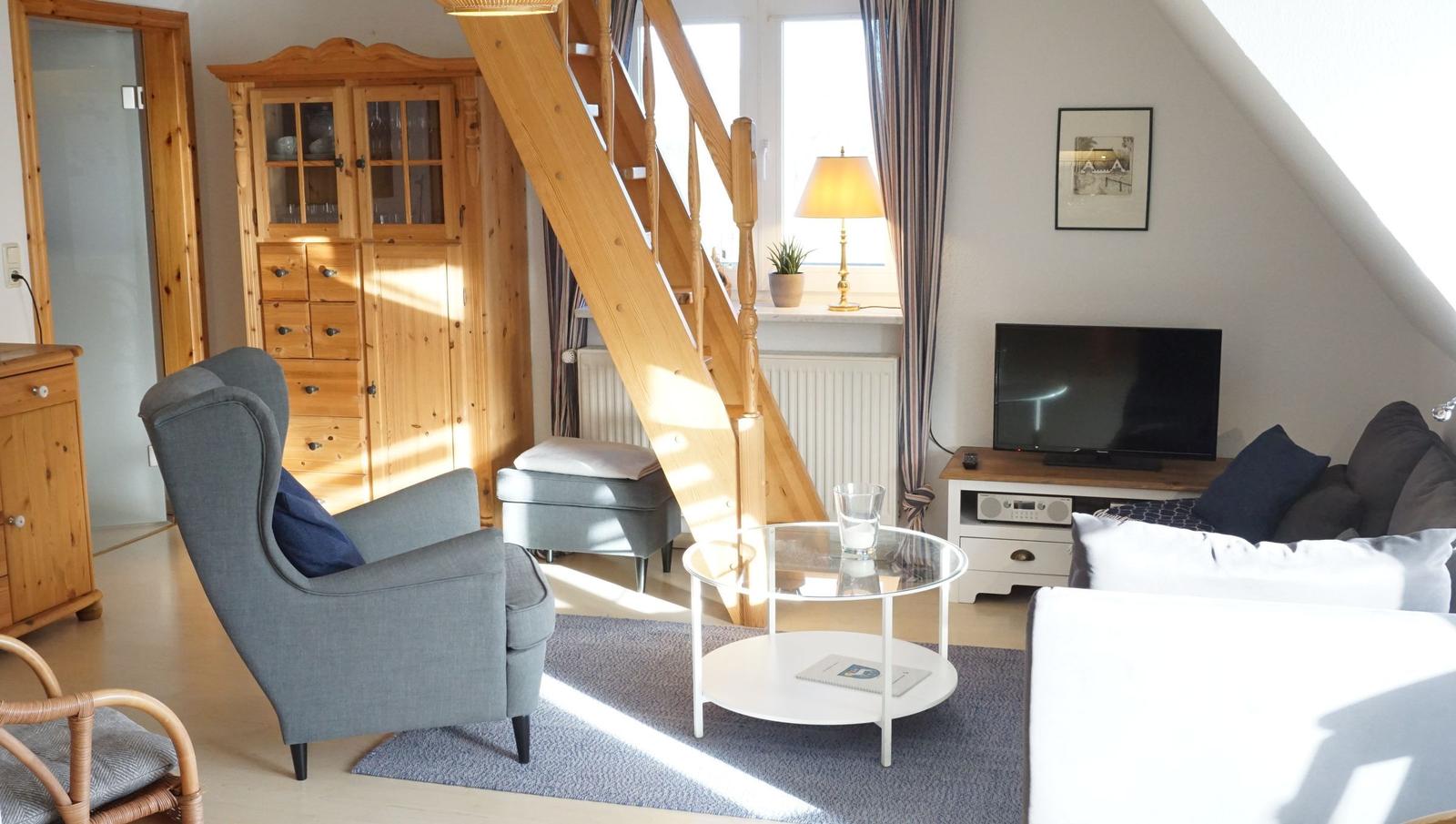 Wohnzimmer mit Sitzgruppe, Glas-Tisch und Holztreppe. Fernseher, Schrank und Fenster sichtbar.