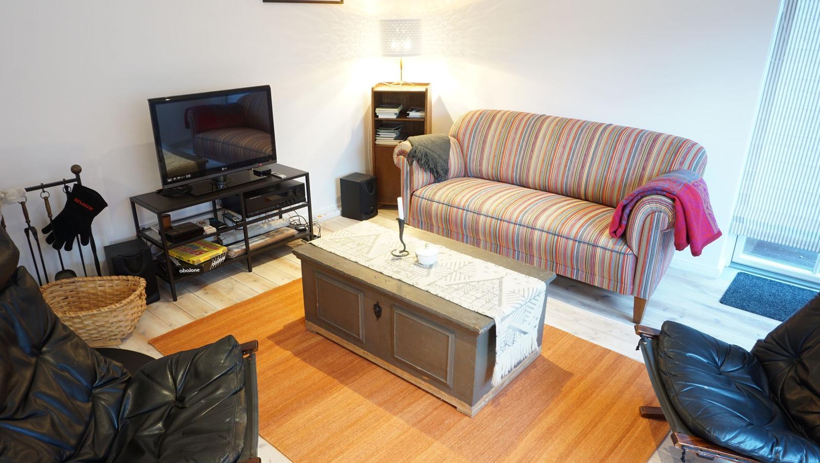 Ein Wohnzimmer mit Sofa, Fernseher und Holztisch. Zwei Ledersessel und eine Tür sind sichtbar.