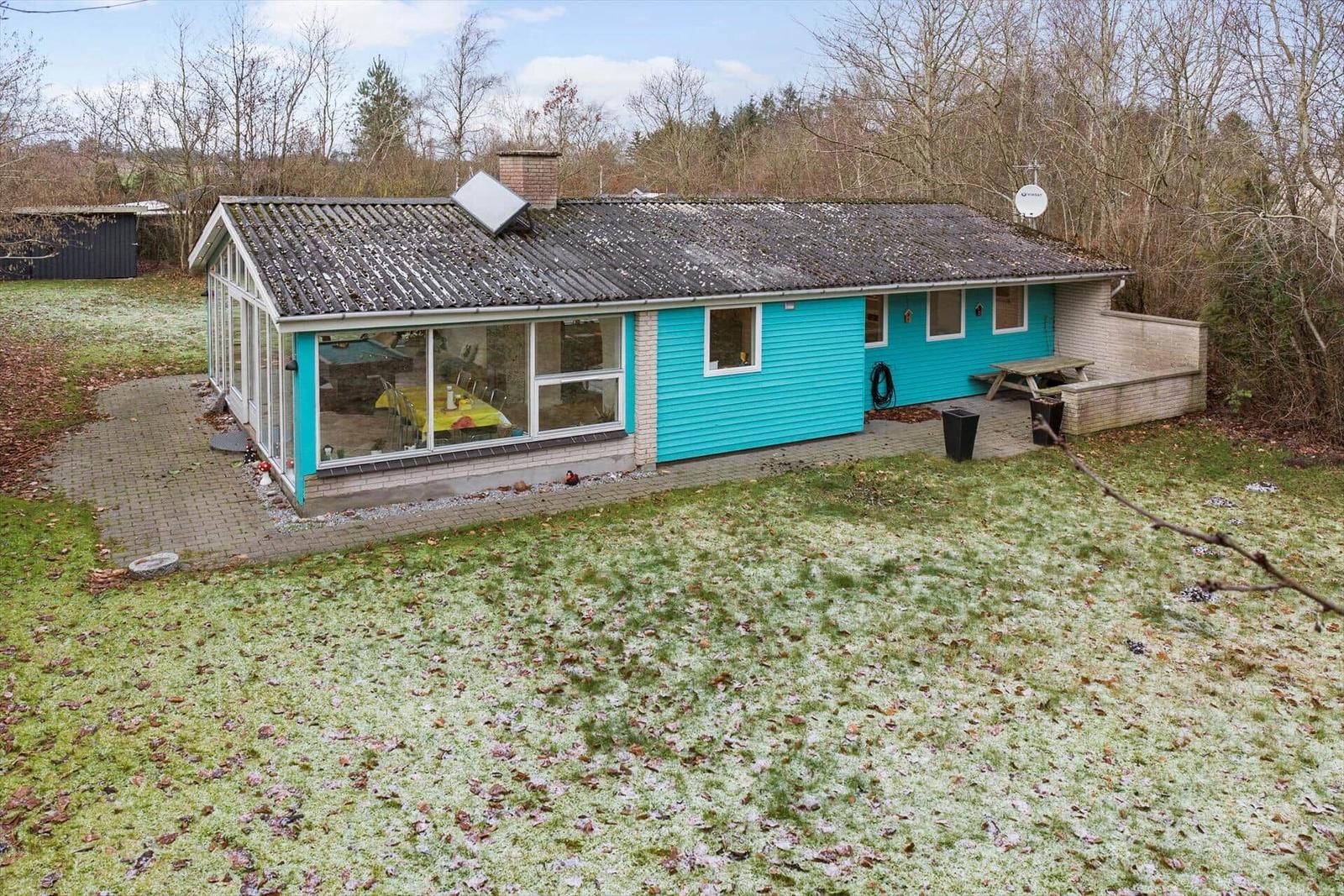 Ein blaues Haus mit Glasfront und Terrasse im Garten. Gras mit Schnee und Bäume im Hintergrund.