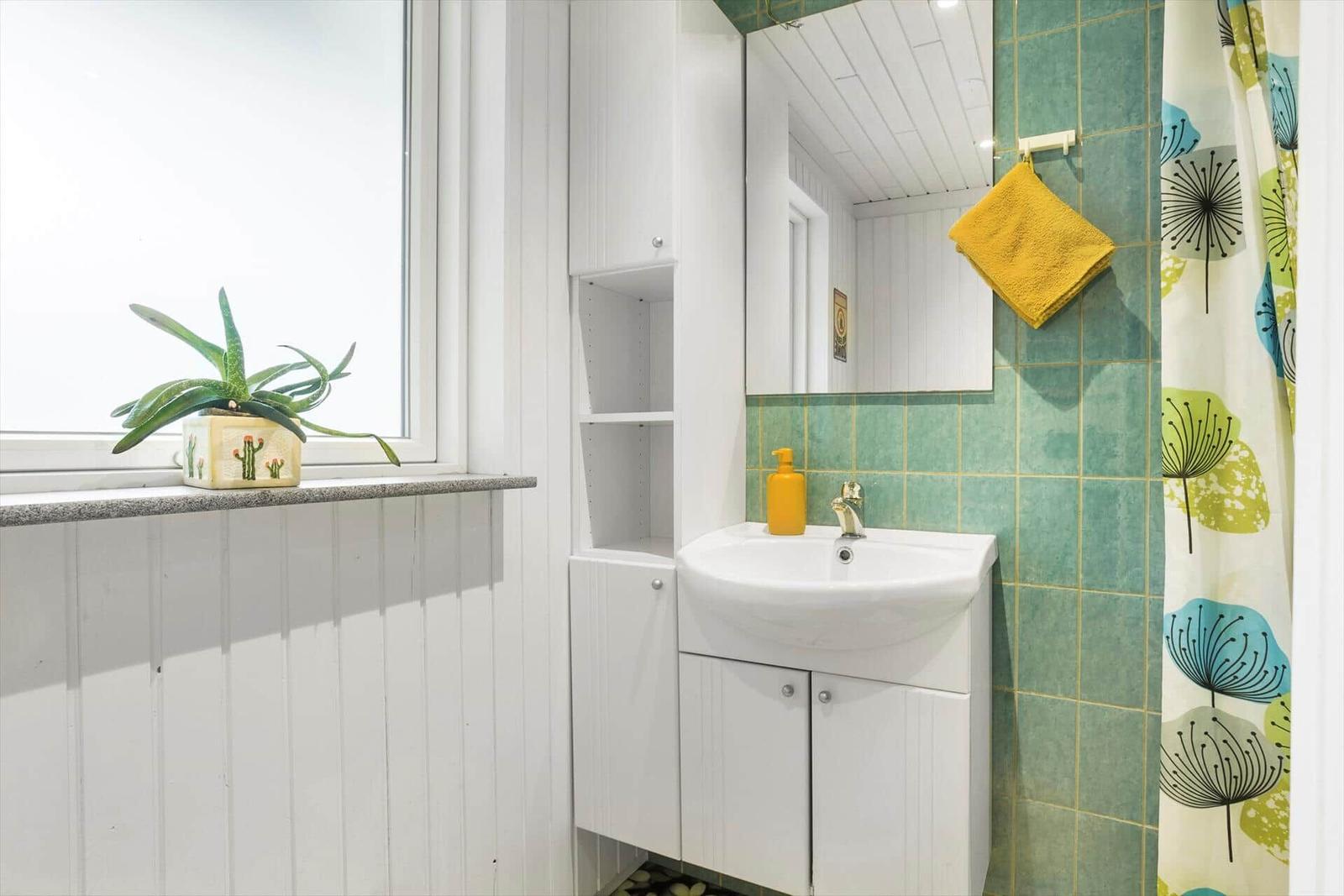 Badezimmer mit Waschbecken, Spiegel, Duschvorhang und Pflanze am Fenster.