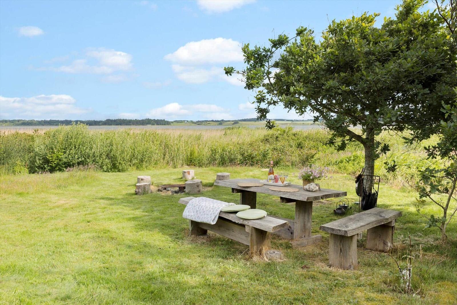 Holztisch mit Bänken unter Baum, Blick auf Grasland und Wasser.