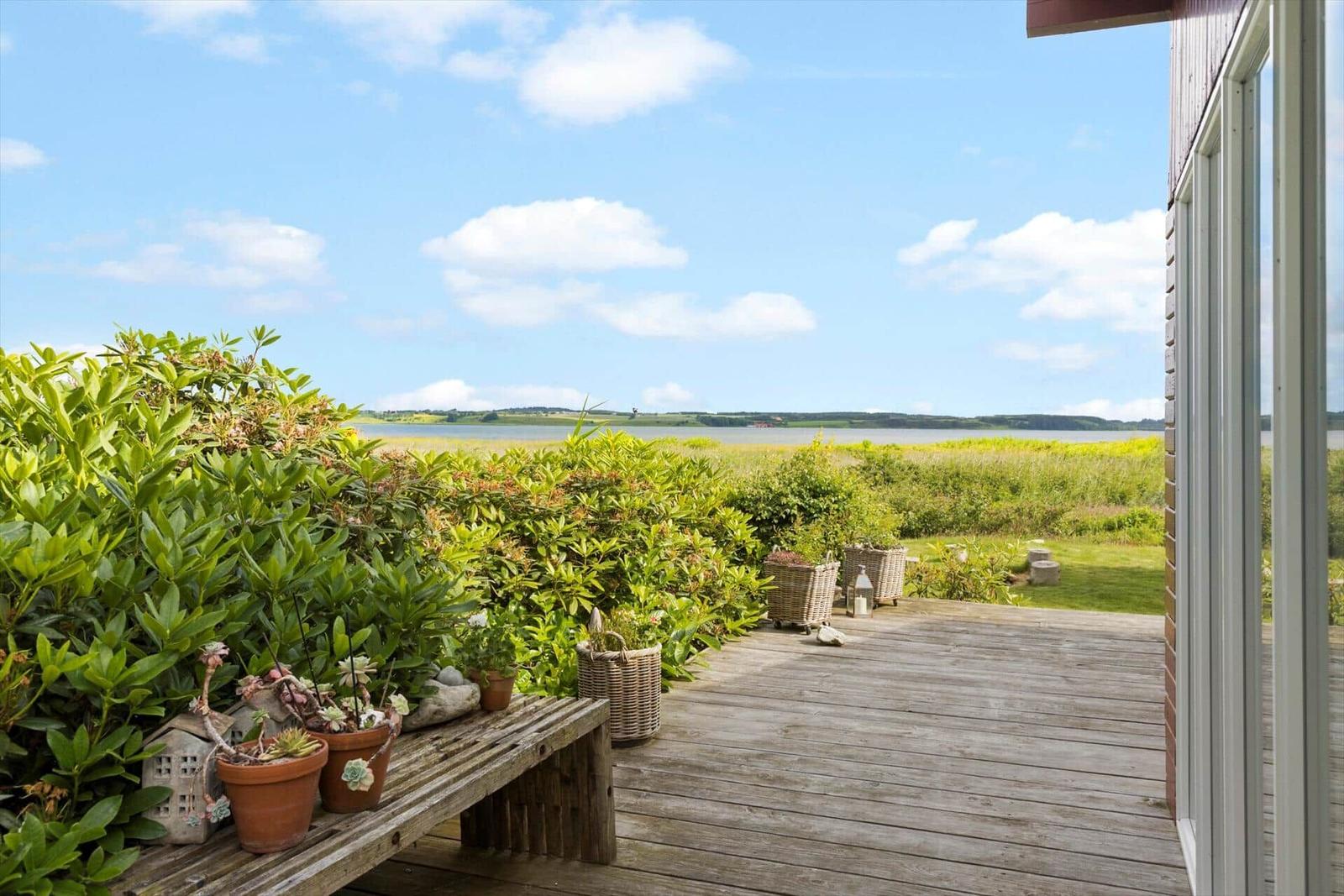 Terrasse mit Blick auf Wasser und Grünflächen. Holzbank, Pflanzen und Körbe sind sichtbar.