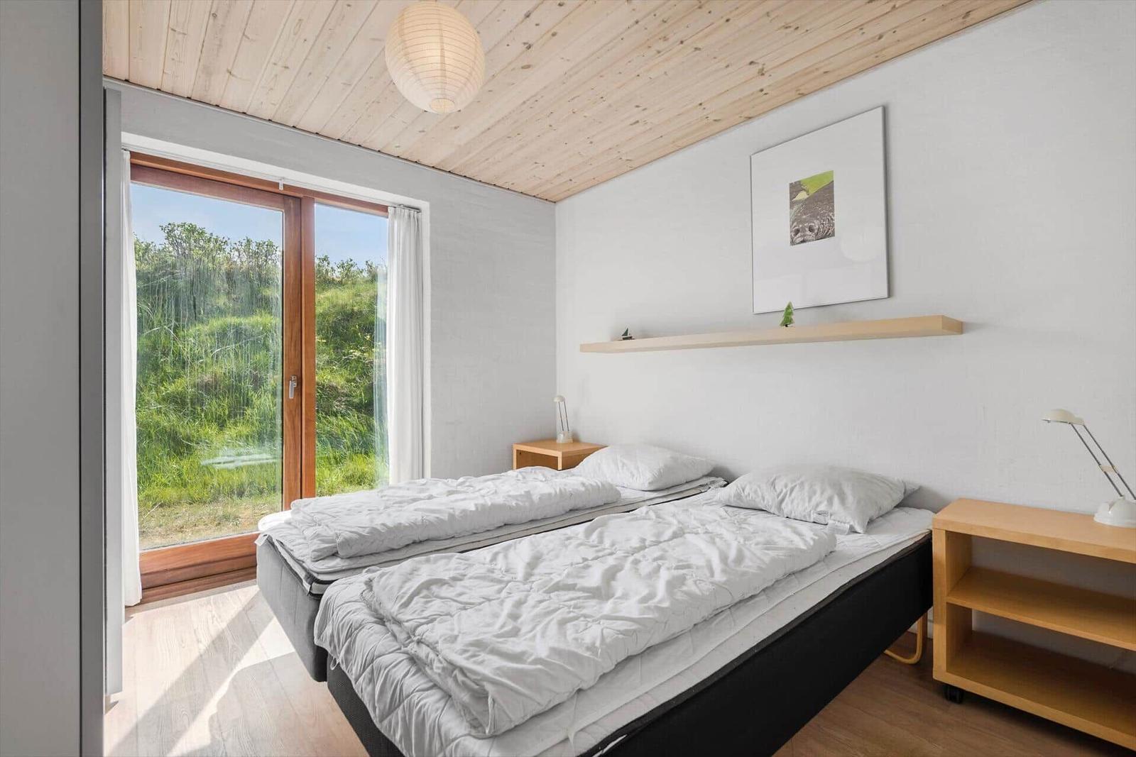 Schlafzimmer mit Doppelbett, Holzdecke und großem Fenster mit Blick auf Grünflächen.