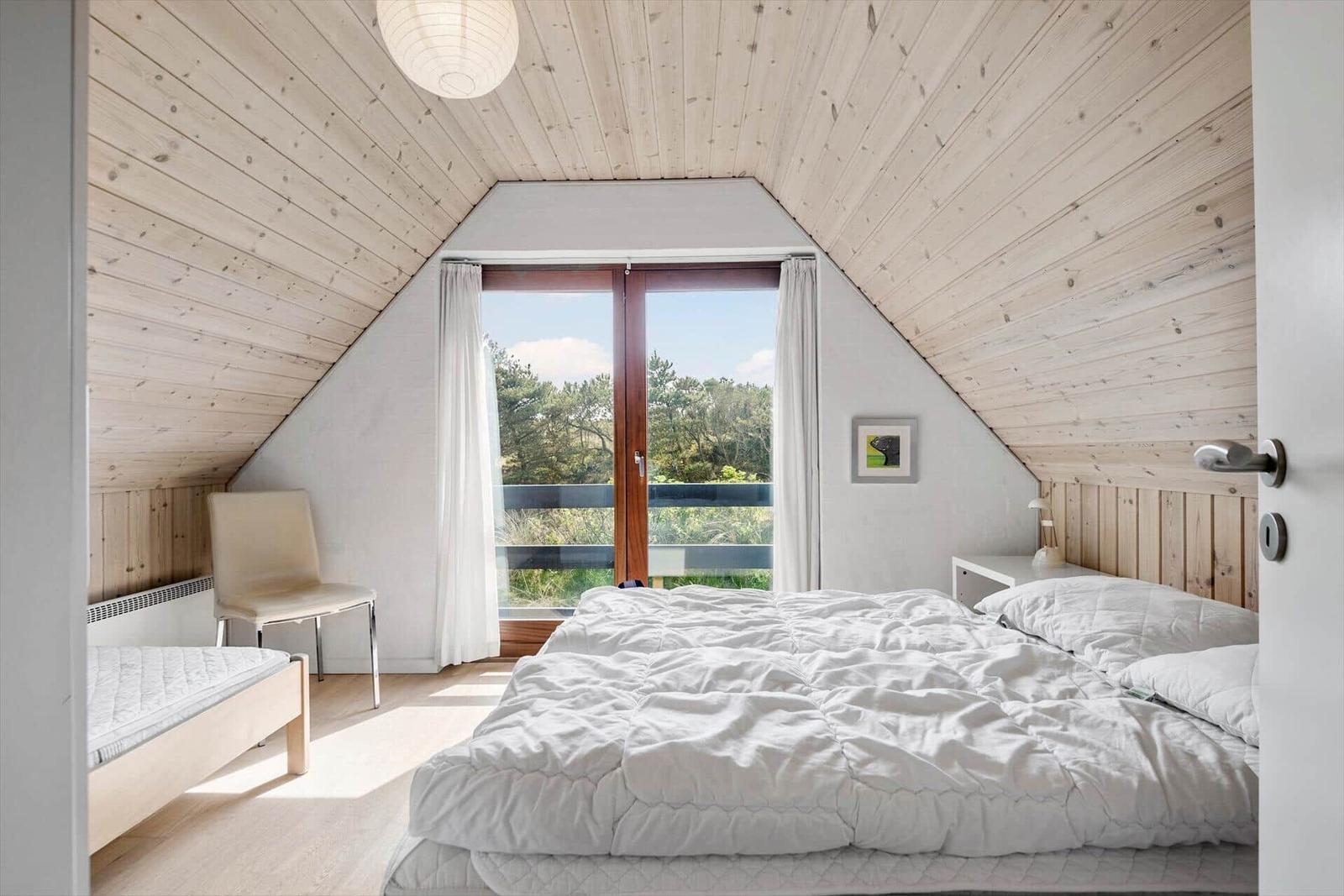 Schlafzimmer mit Dachschrägen, großem Fenster und weißem Bett. Holzwände und Decke.