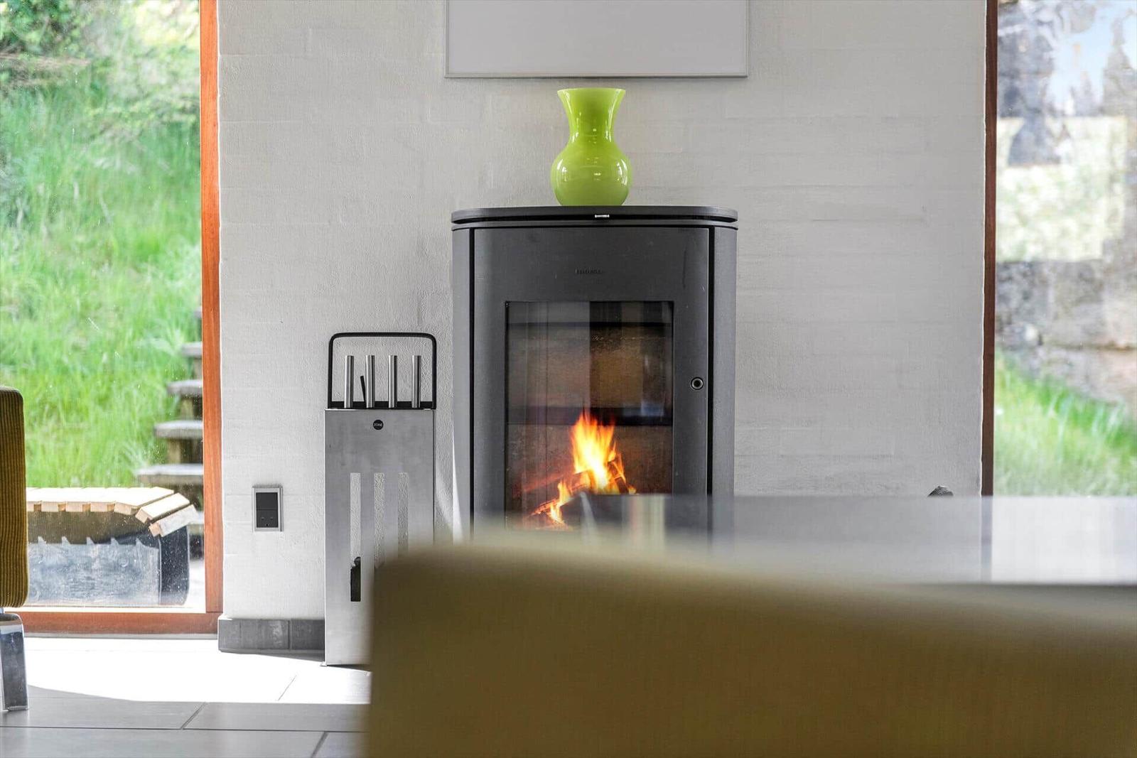 Ein Kamin mit brennendem Feuer steht in einem Raum mit großen Fenstern und einem grünen Vasenobjekt darauf.