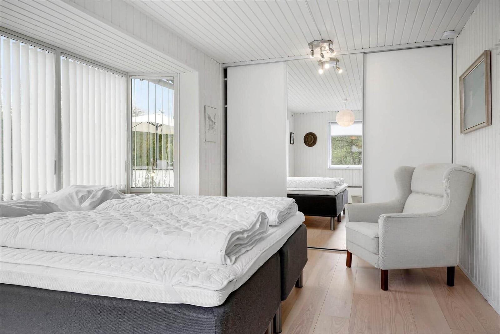 Schlafzimmer mit Bett, Sessel und Fenster. Spiegelwand zeigt weiteren Raum.