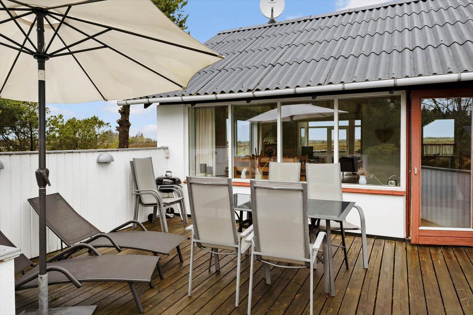 Terrasse mit Sonnenschirm, Liegestühlen und Tisch. Blick in das Haus mit Glasfront.