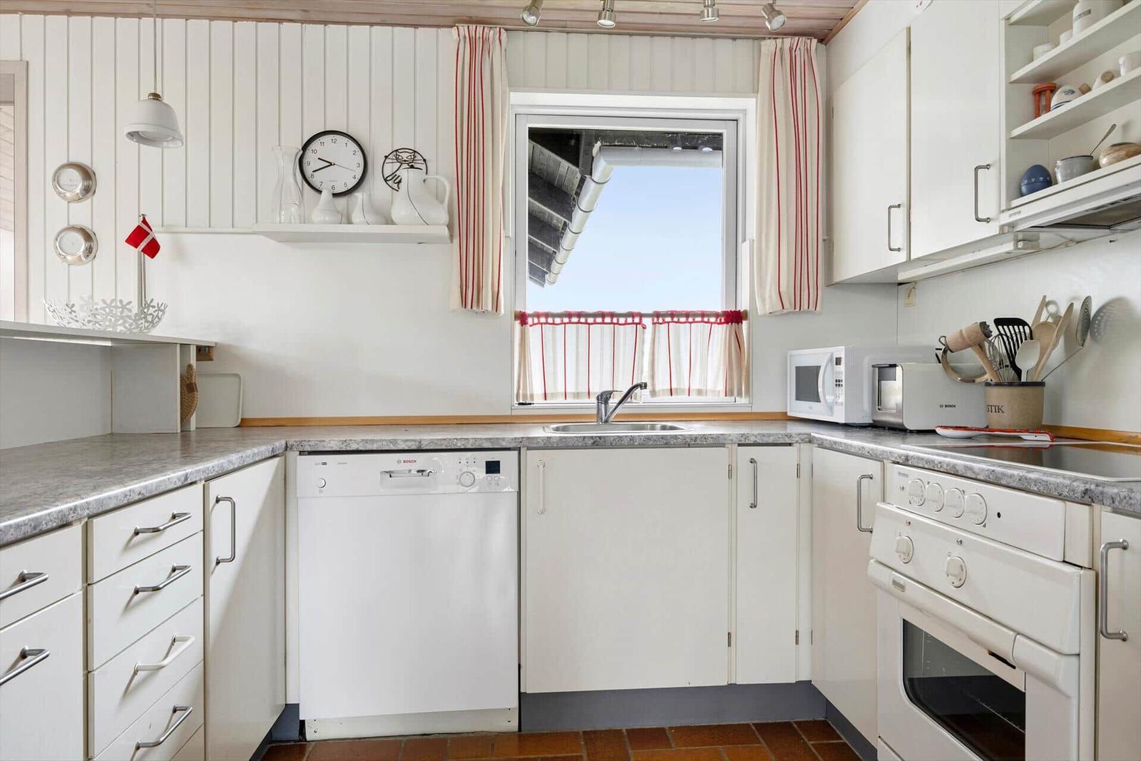 Küche mit weißen Schränken, Spüle, Herd und Waschmaschine. Fenster mit roten Vorhängen.