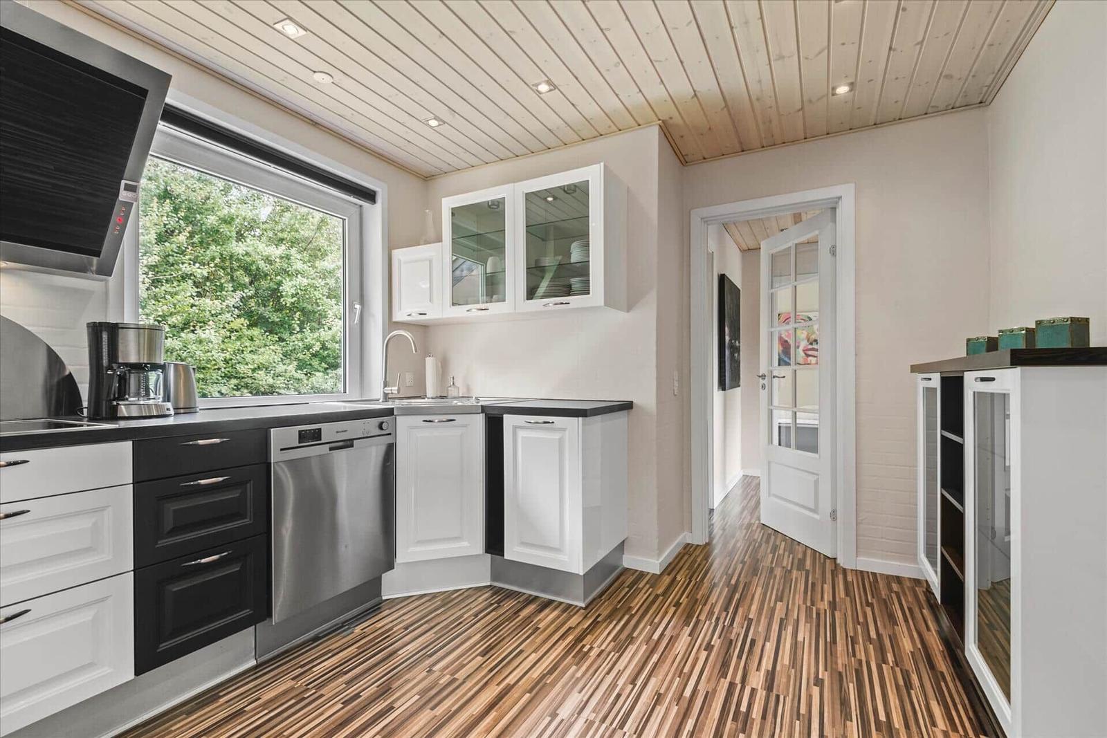 Küche mit weißen Schränken, dunklem Arbeitsplatz und Holzboden. Fenster mit Blick auf Bäume.