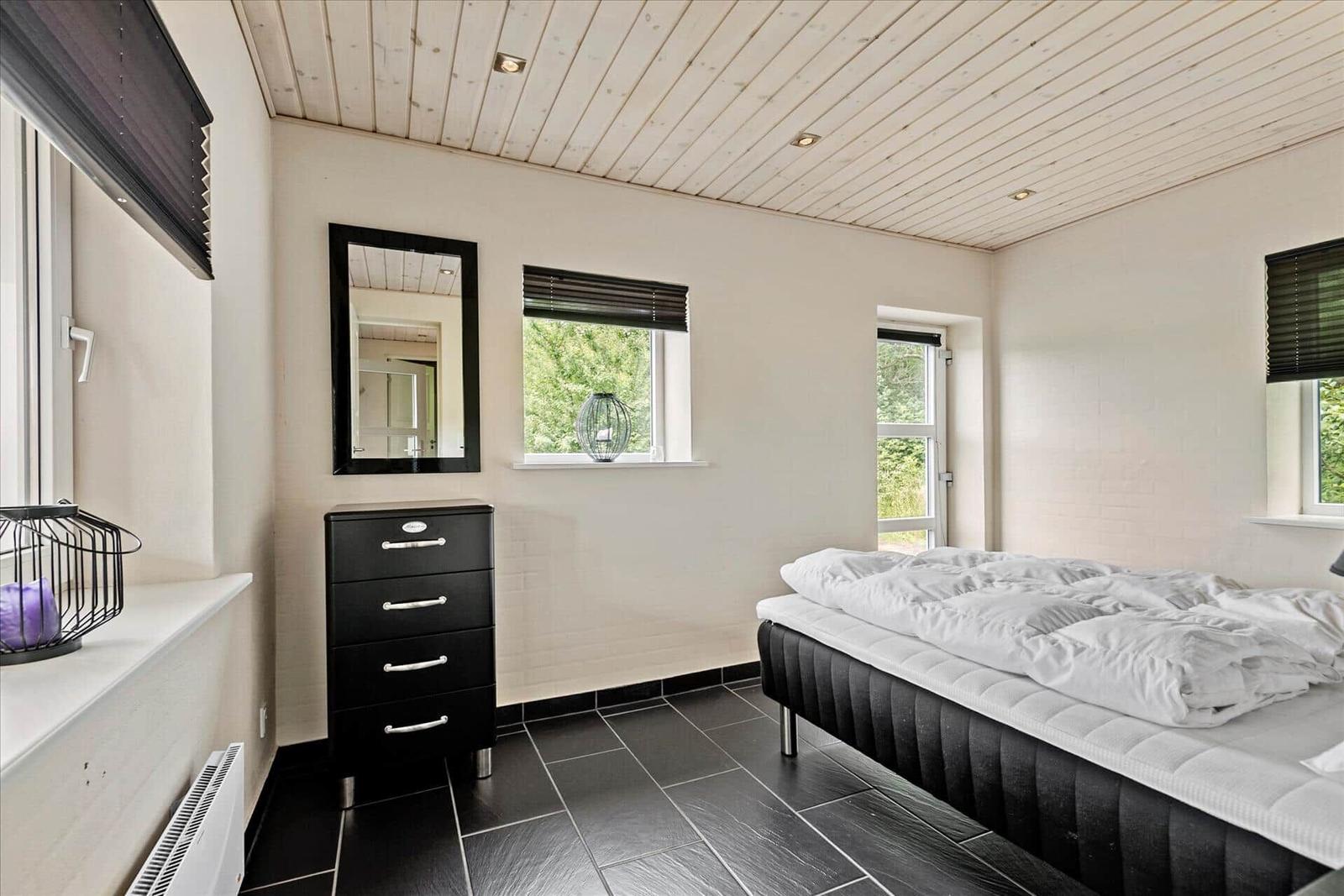 Schlafzimmer mit Bett, Schrank und Fenstern. Holzdecke und dunkle Fliesen.