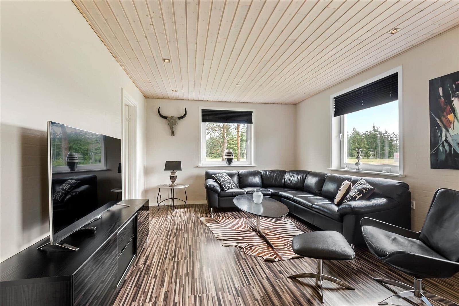 Wohnzimmer mit schwarzer Ledercouch, Holzboden und TV. Fenster zeigen Wald.