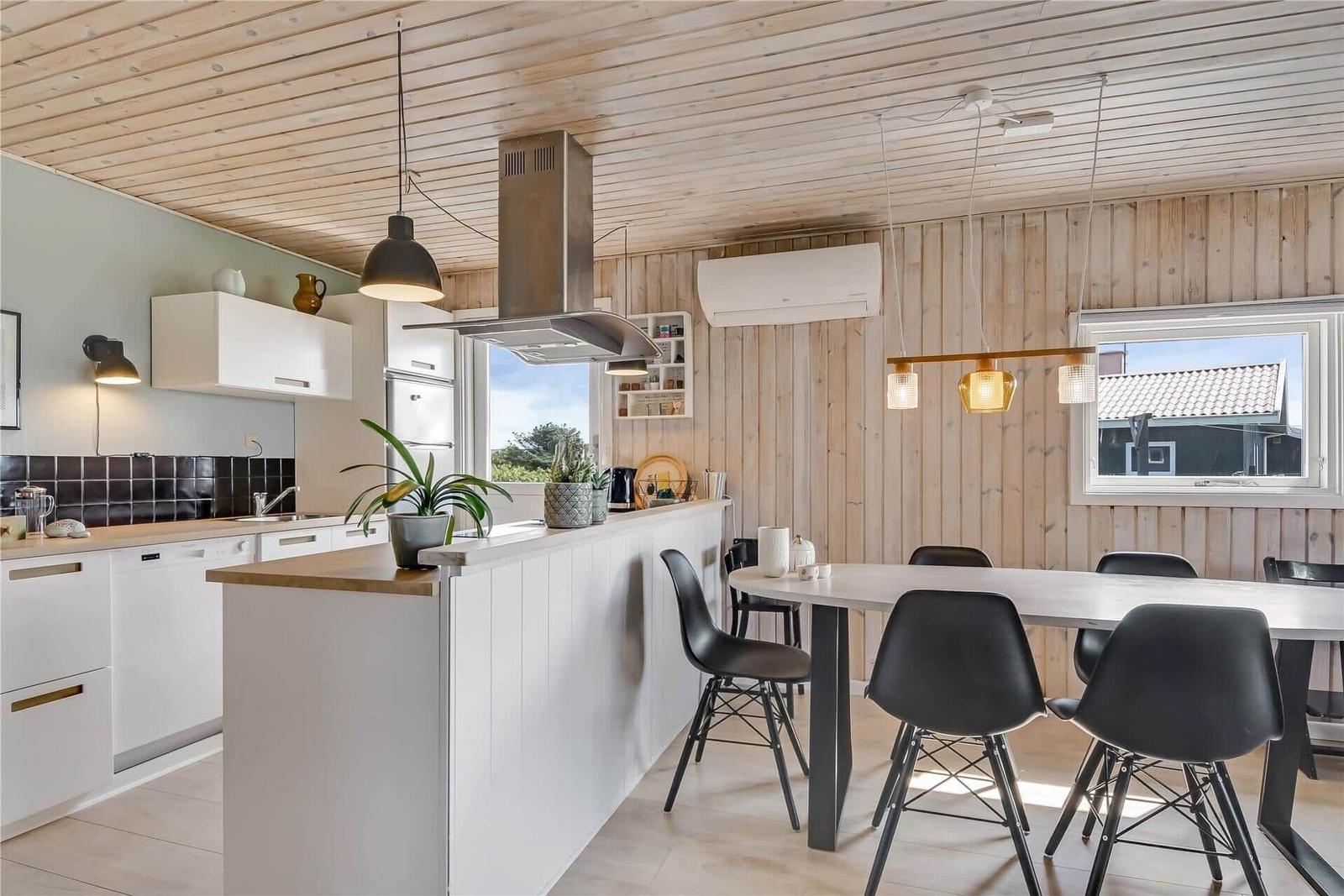 Küche mit Essbereich, Holzwand und -decke, weiße Schränke, schwarze Stühle und Fenster.