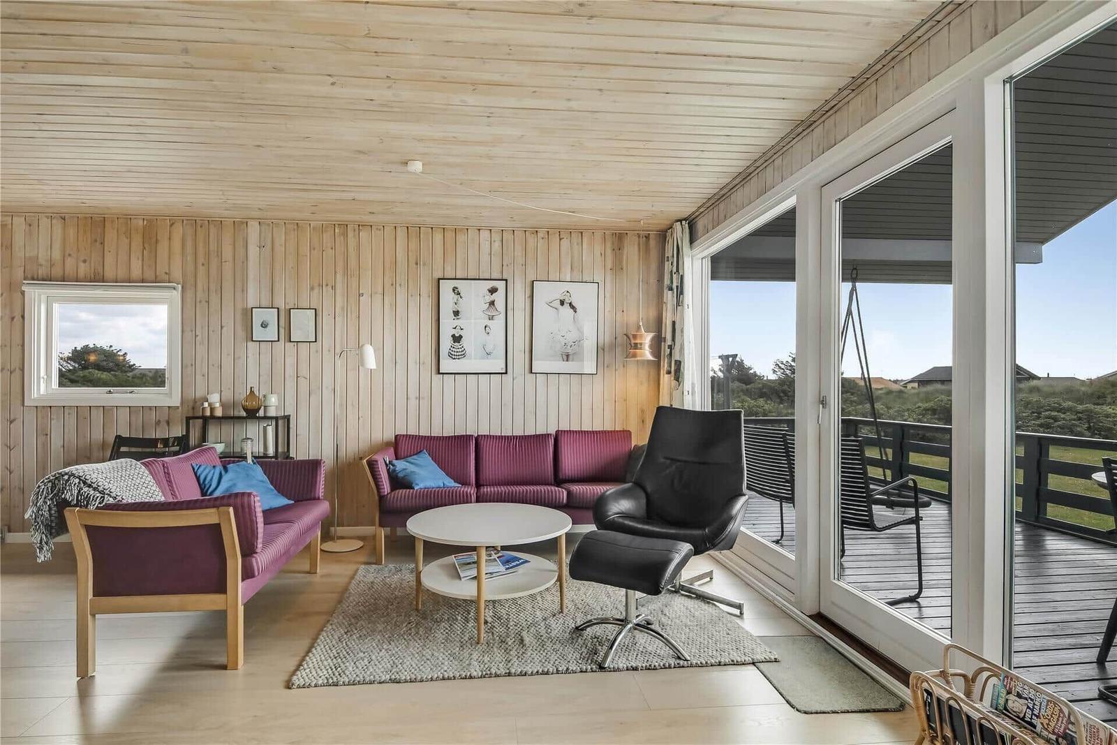 Wohnzimmer mit Holzwand, zwei Sofa, Tisch und Sessel. Durchgang zur Terrasse mit Blick auf Grünflächen.