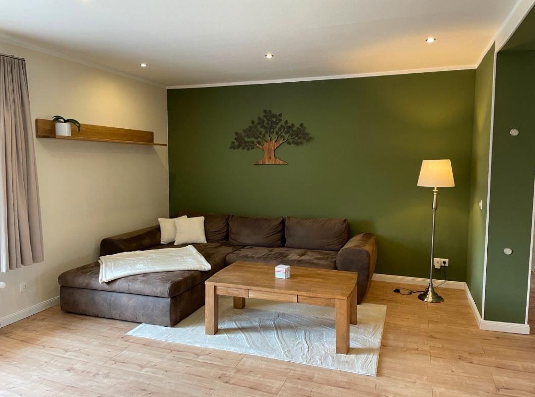 Wohnzimmer mit braunem Sofa, Holztisch und grüner Wand mit Baumdekoration.