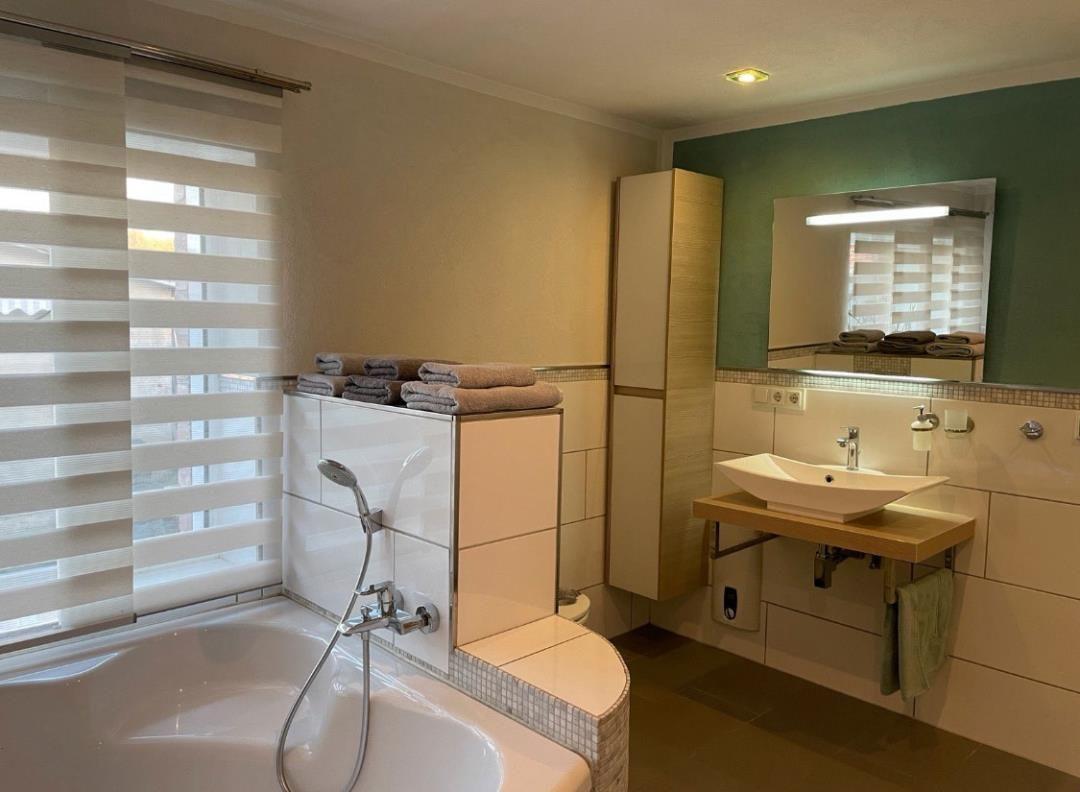 Badezimmer mit Badewanne, Waschbecken und Spiegel. Fenster mit Jalousie.