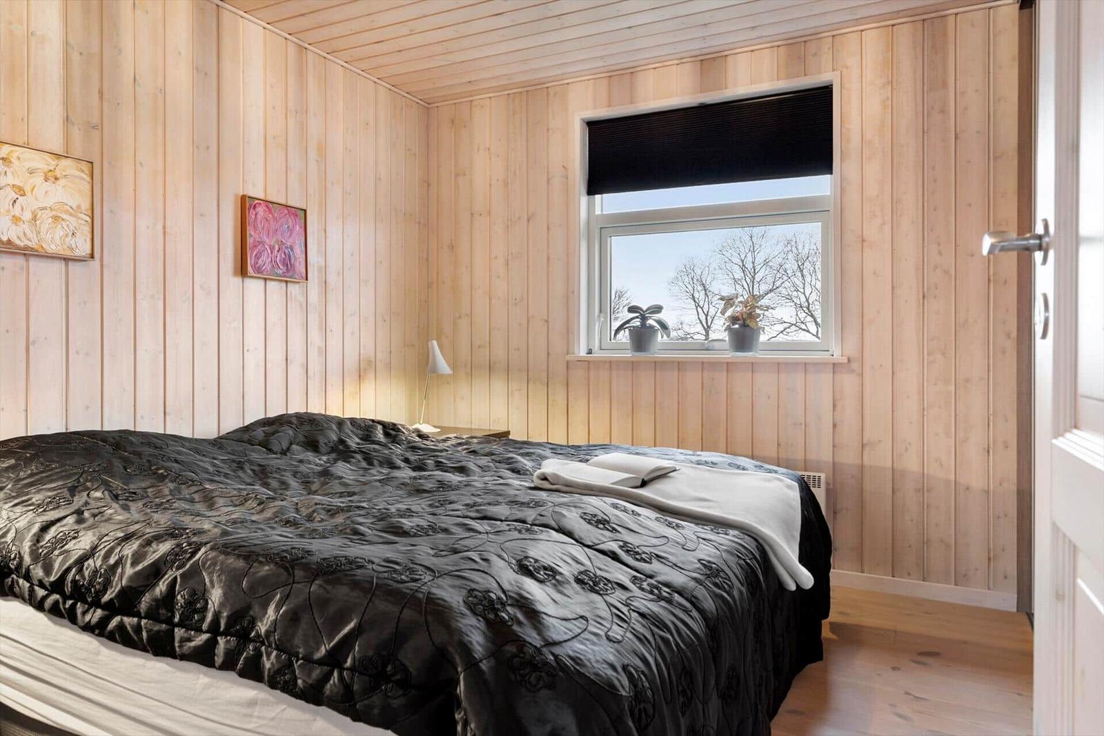 Schlafzimmer mit Holzwänden, Bett mit dunkler Bettwäsche und Fenster mit Blick auf Bäume.