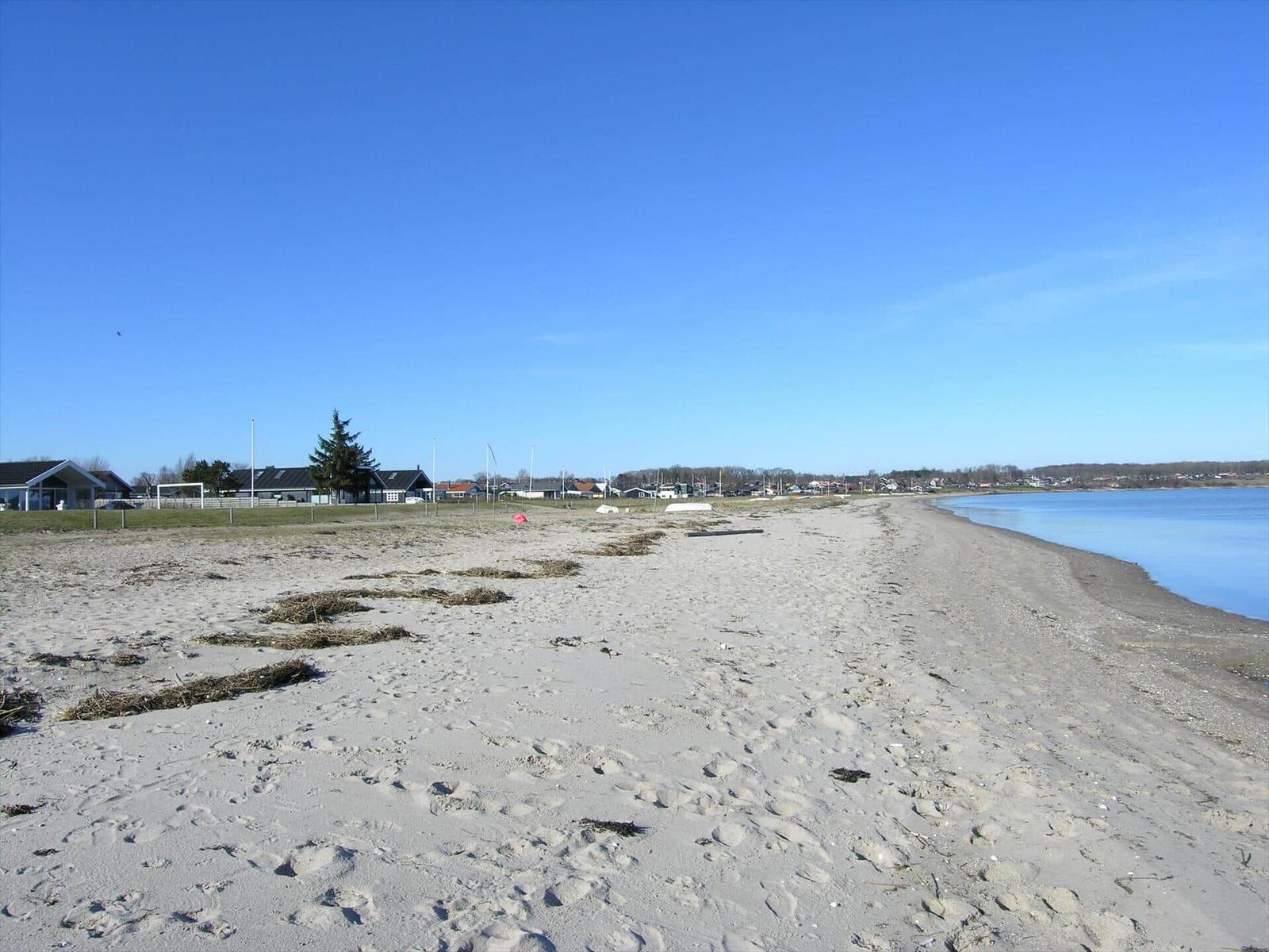 Strand mit Sand und Wasser, im Hintergrund Häuser und Bäume unter blauem Himmel.