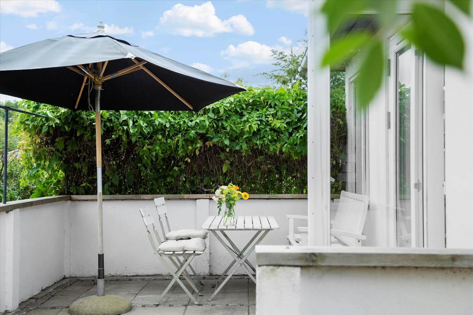 Terrasse mit Tisch, Stühlen und Sonnenschirm. Hintergrund: grüner Heckenzaun.