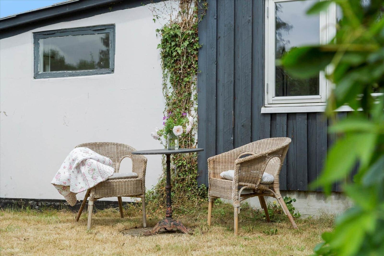 Zwei Rattan-Stühle und ein Tisch stehen im Garten vor einem Haus mit dunklem Holz und weißer Wand.