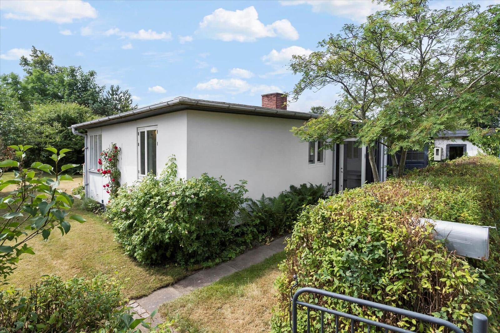 Ein weißes Haus mit Garten, Fenstern und einem Schornstein. Umgeben von Bäumen und Sträuchern.
