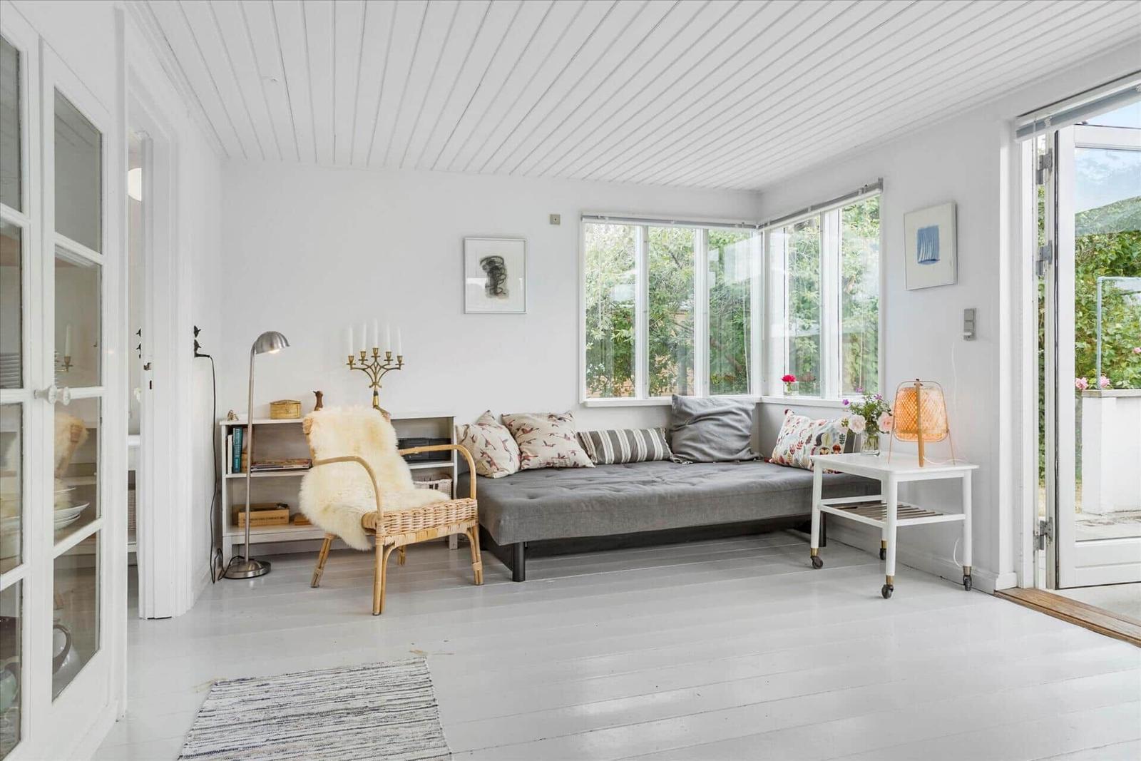 Wohnzimmer mit grauem Sofa, Wickerstuhl und großen Fenstern. Weiße Wände und Holzboden.