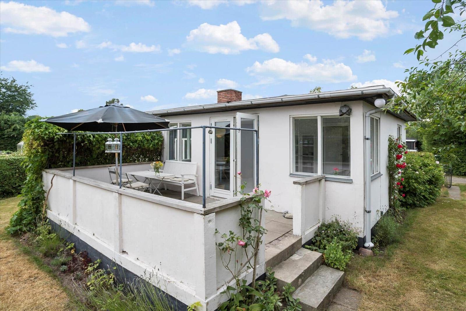 Weiße Villa mit Terrasse, Sonnenschirm und Garten. Eingangstür und Fenster sichtbar.