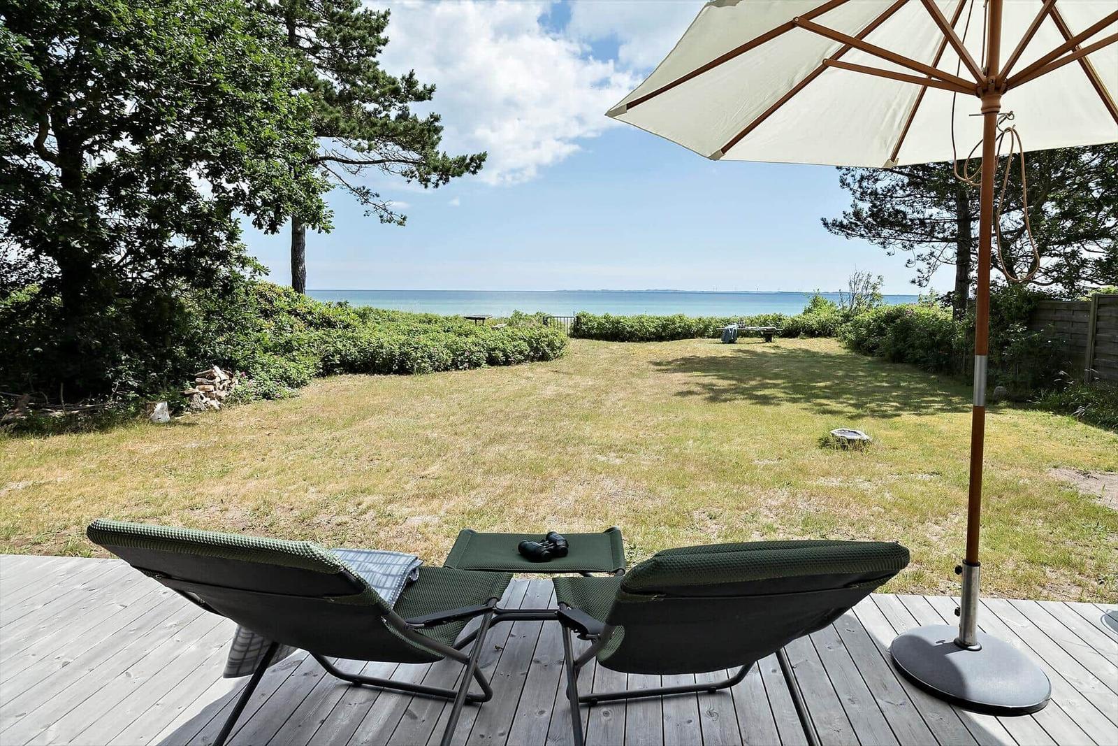 Terrasse mit Liegestühlen und Sonnenschirm. Blick auf Grasfläche und Meer.