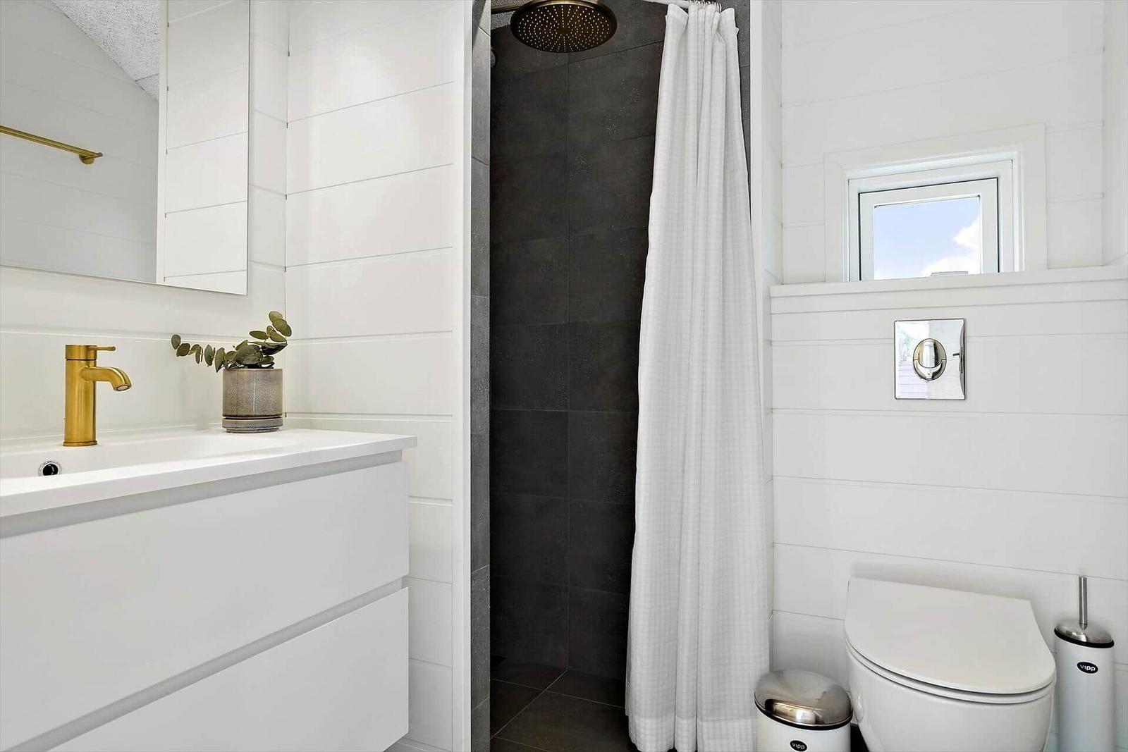 Badezimmer mit Dusche, Waschbecken und Toilette. Weiße Wände und goldener Wasserhahn.