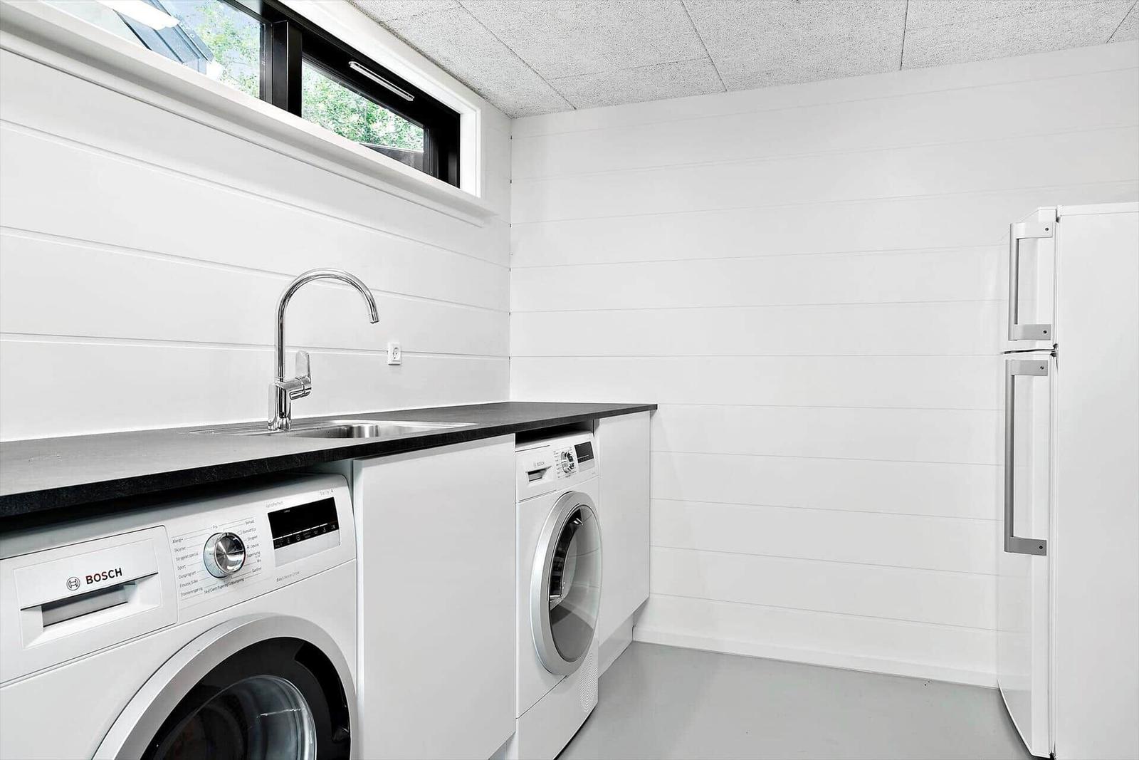 Waschraum mit Waschmaschine, Spüle und Kühlschrank. Weiße Wände und schwarzer Arbeitsplatz.