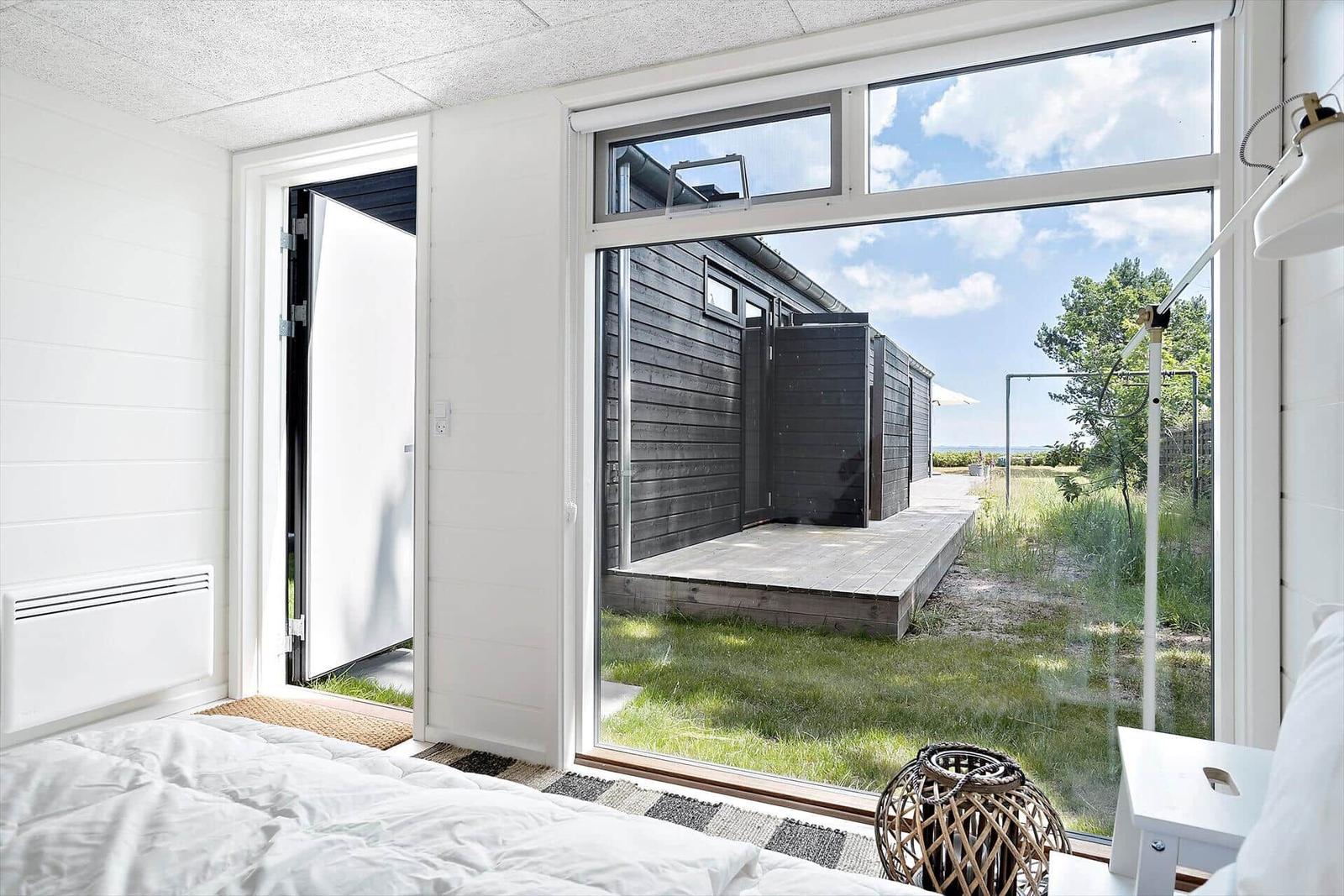 Schlafzimmer mit Blick auf Holzterrasse und grünen Garten. Weiße Wände und große Fenster.