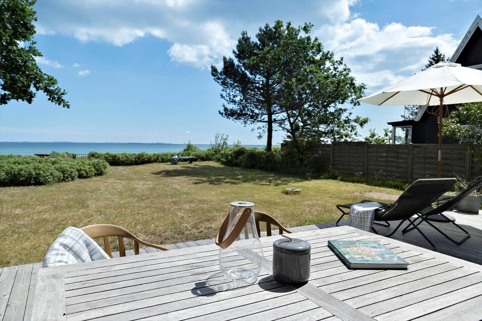 Terrasse mit Tisch, Stühlen und Sonnenschirm. Blick auf Grasfläche und Meer.