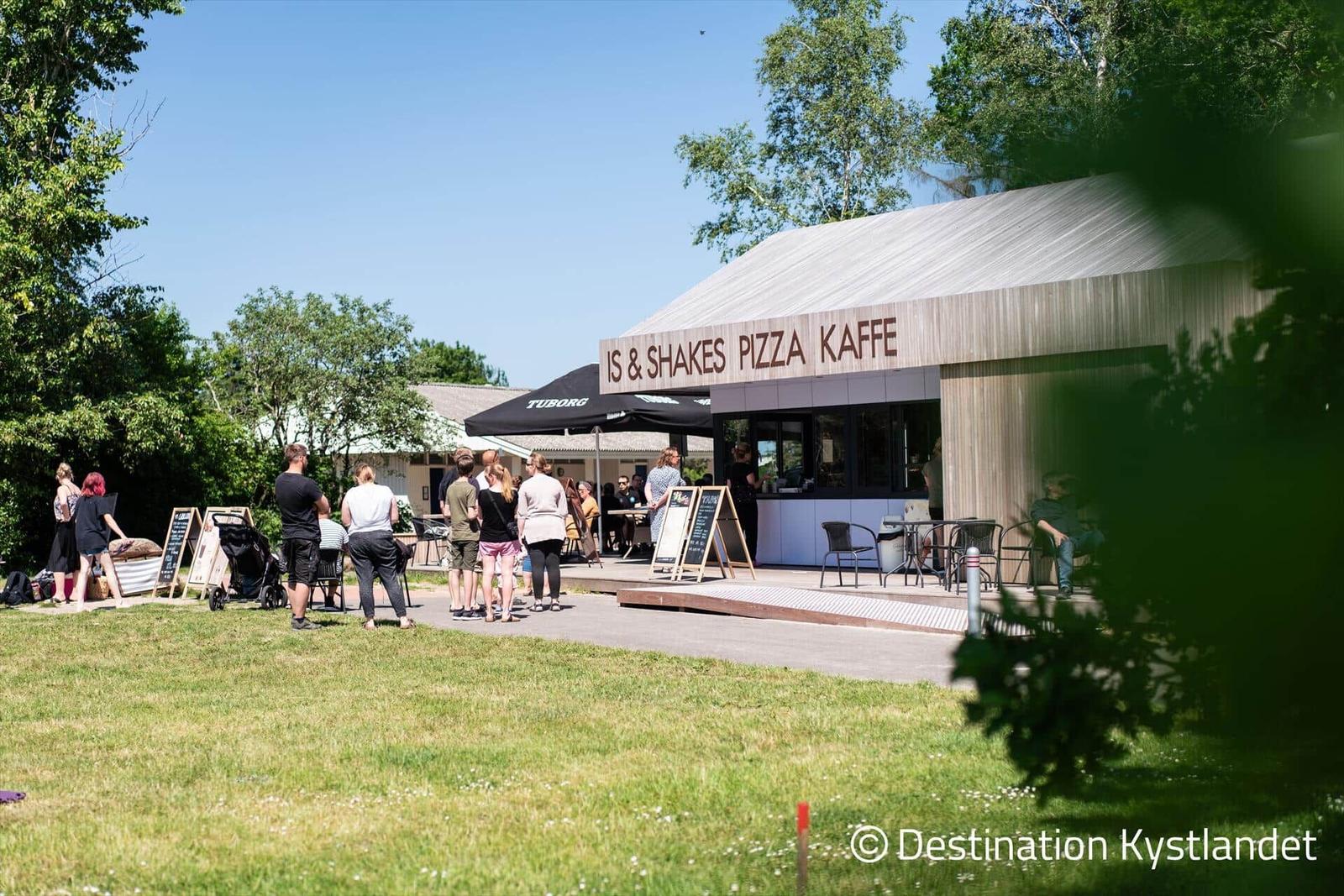 Kaffee- und Pizza-Stand mit Außenterrasse und Gästen unter Sonnenschein.