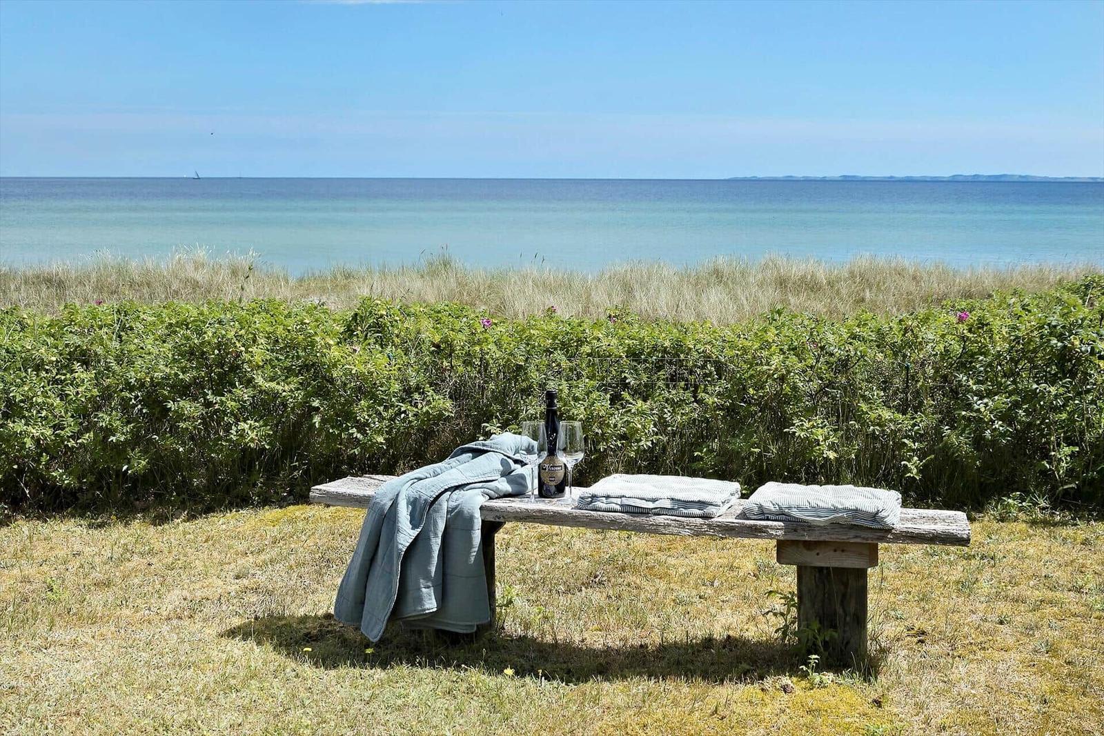 Holzbank mit Decke und Getränk vor Strand und Gras.