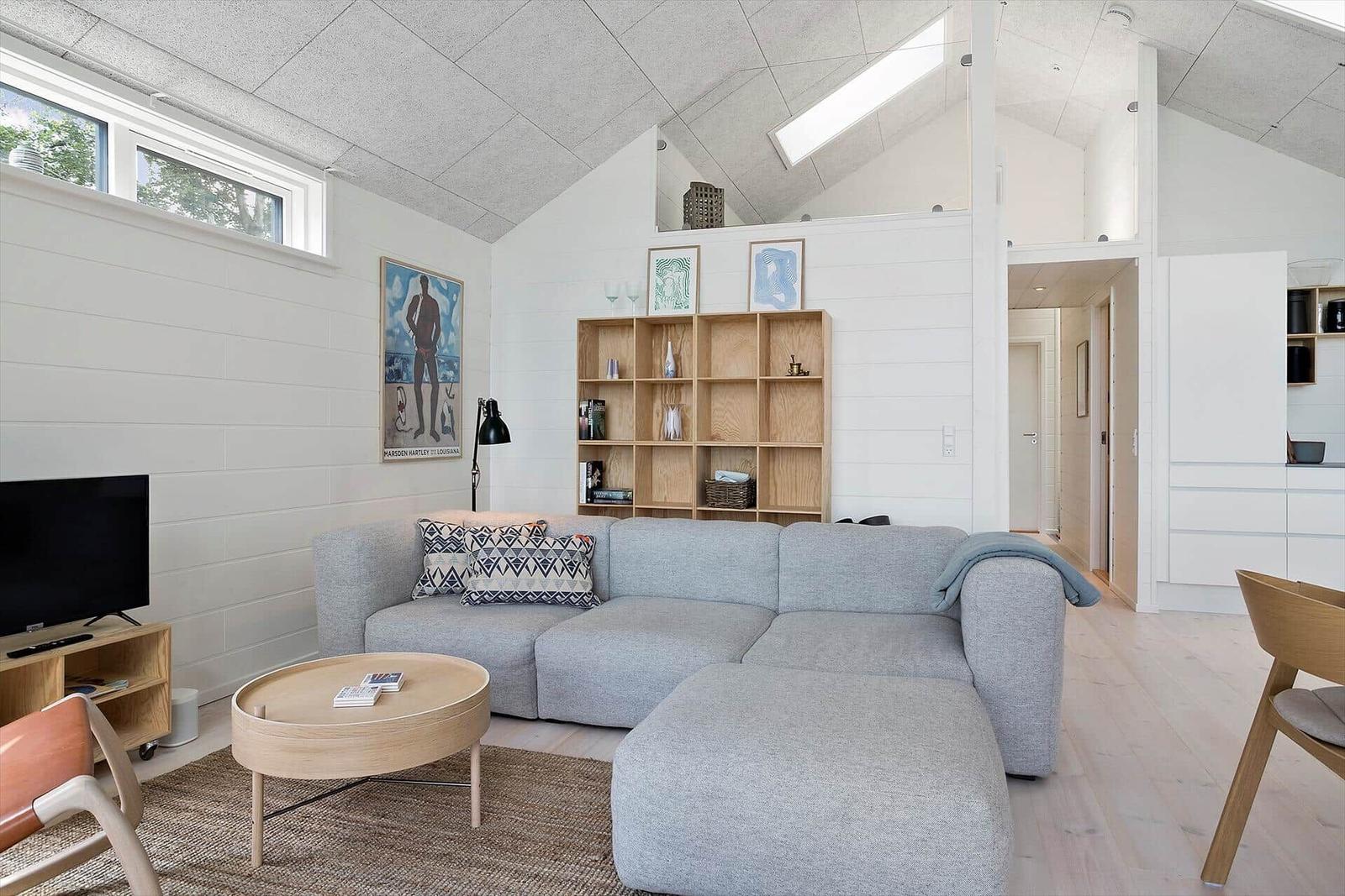 Wohnzimmer mit grauem Sofa, Holzregal und Fernseher. Holzfußboden und weiß gestrichene Wände.