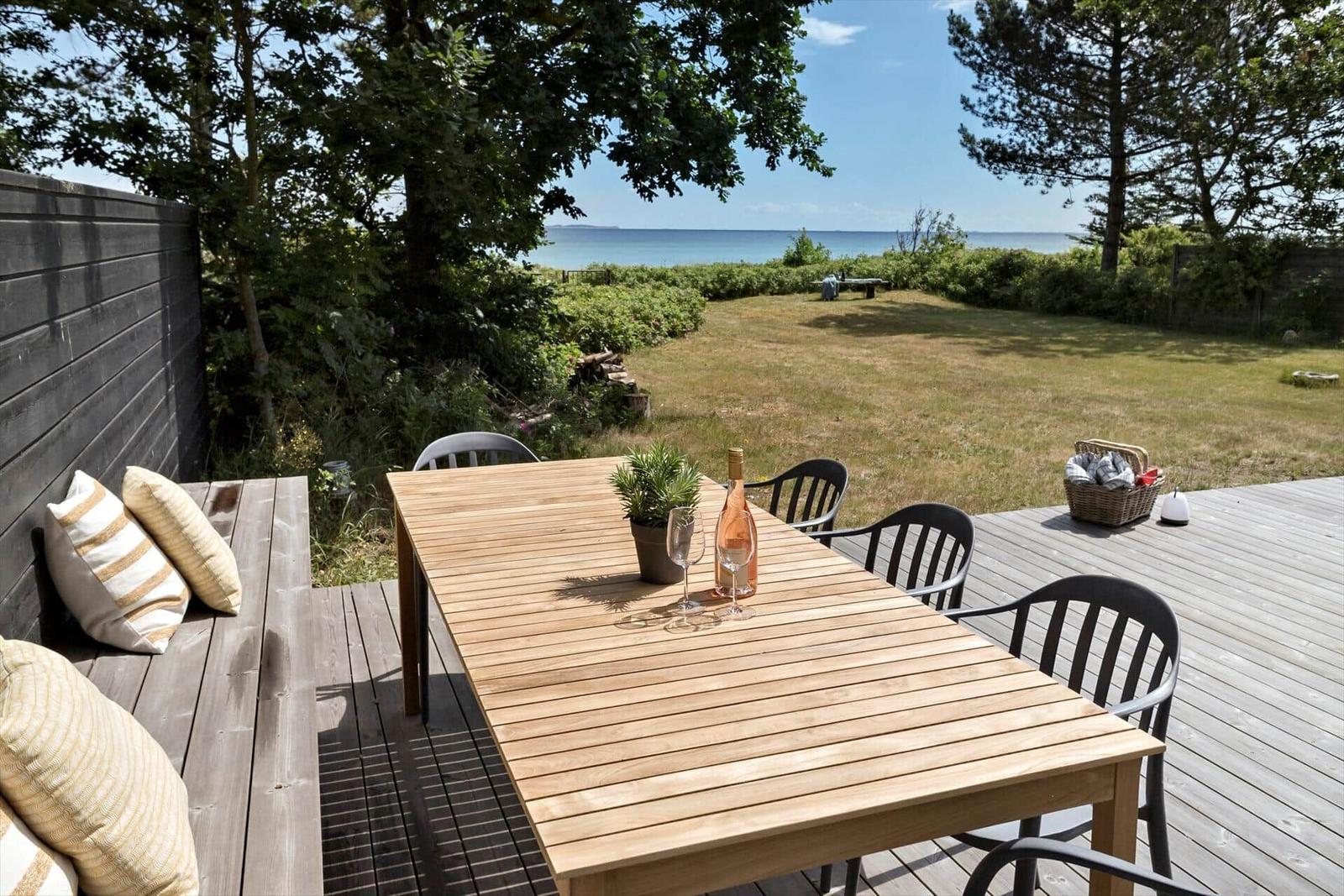 Holztisch mit Stühlen auf Terrasse, Blick auf Grasfläche und Meer.