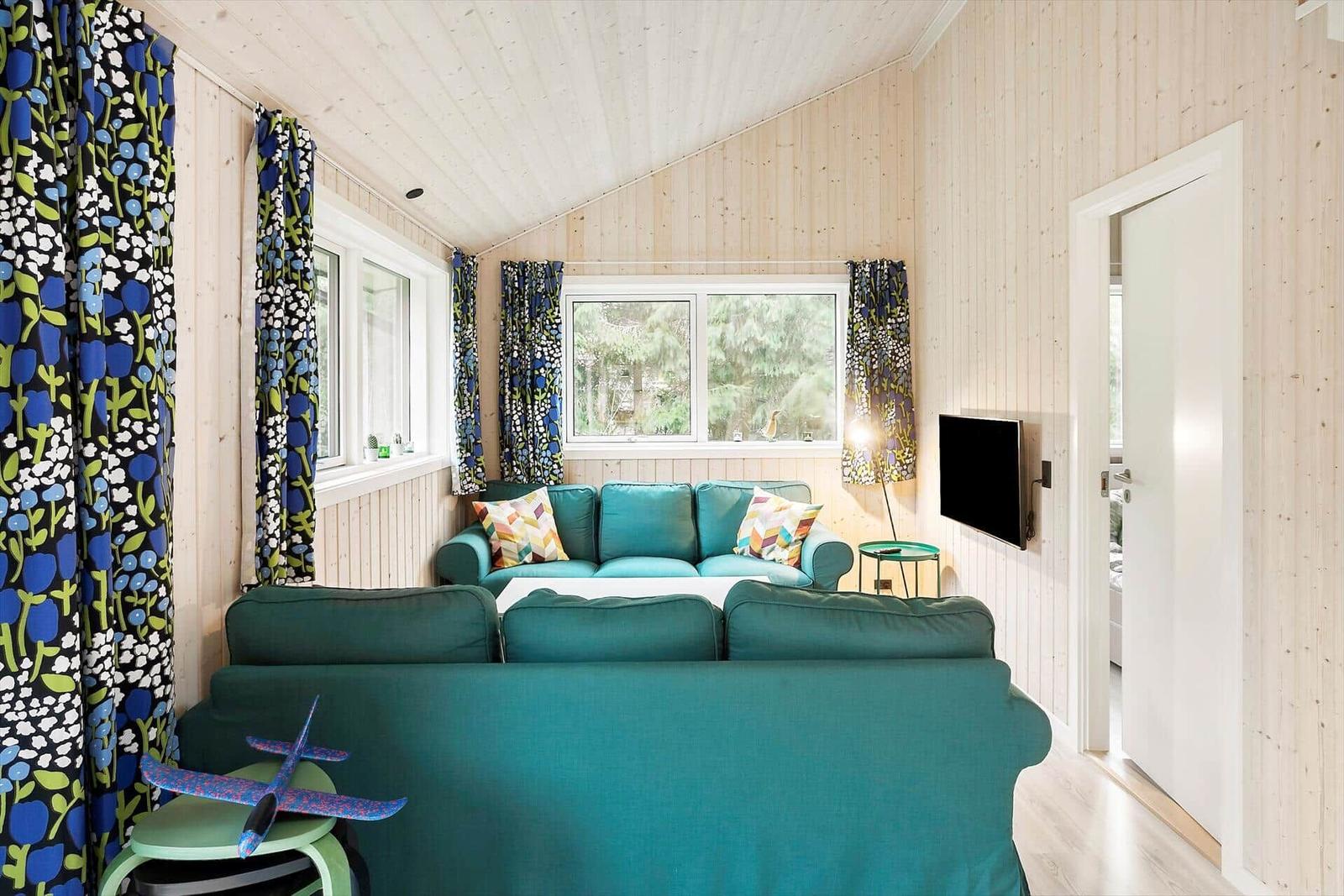 Living room with green sofa, windows, and wooden walls. TV and curtains are visible.