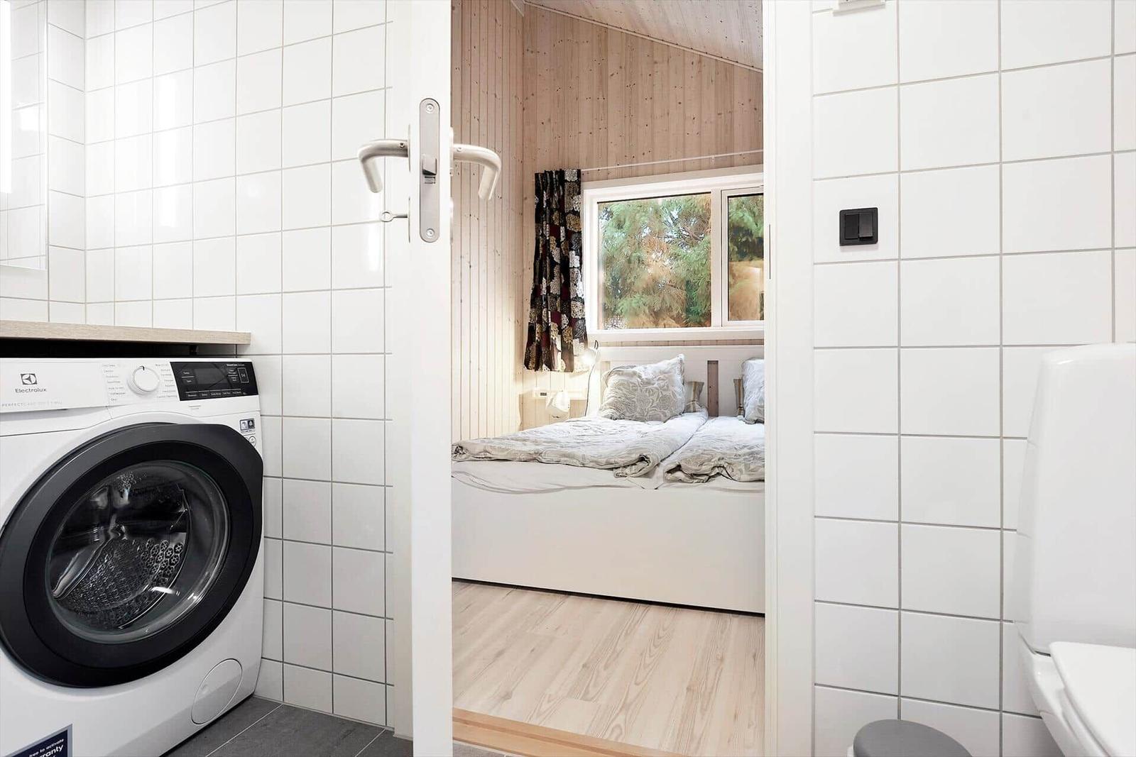 Washing machine in bathroom, view into bedroom with bed and window.